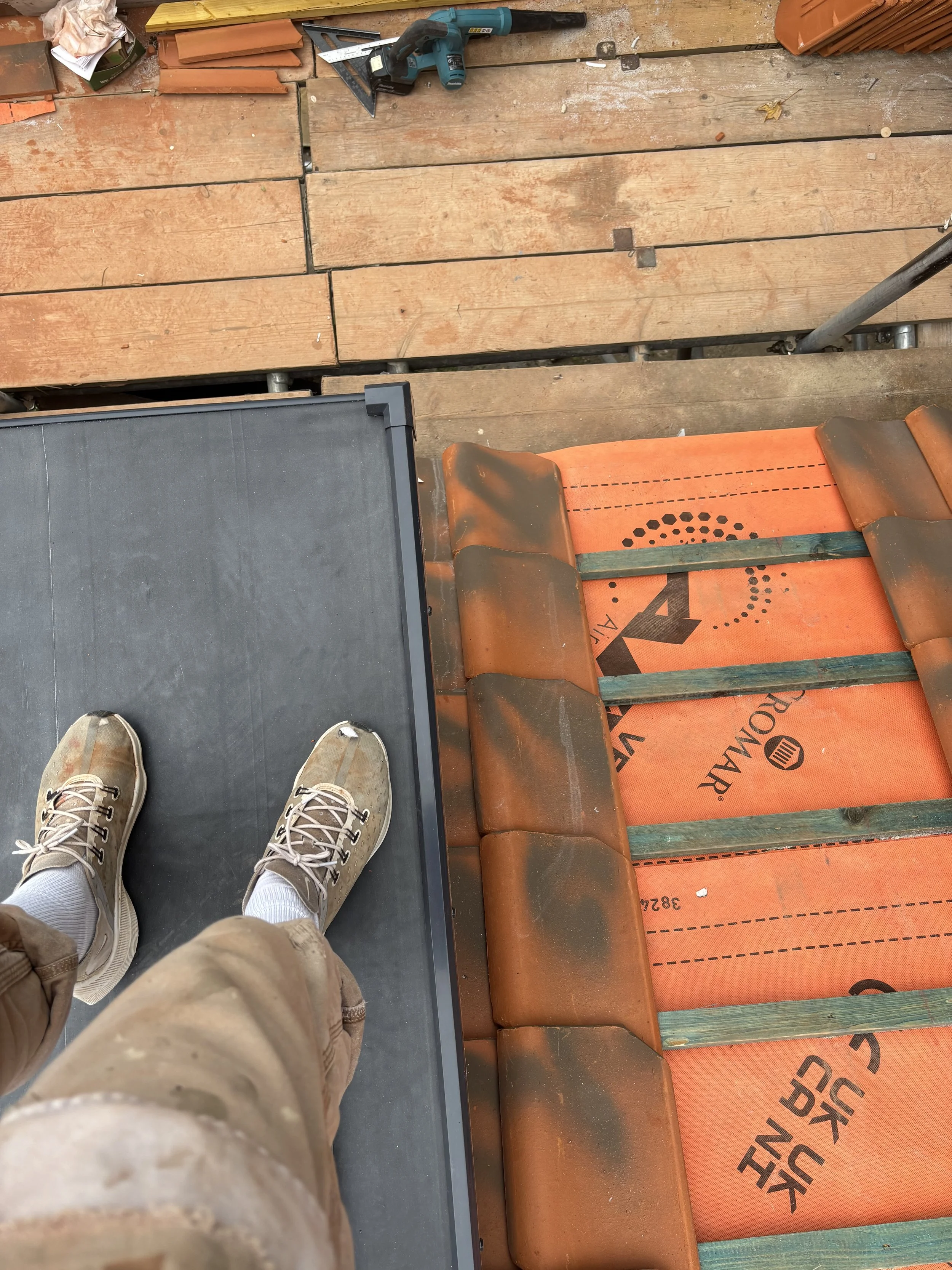View from a person's perspective standing on a construction or renovation site. Person is wearing beige sneakers and khaki pants. The floor consists of wooden planks and orange construction underlayment, with some black non-slip mat visible to the si