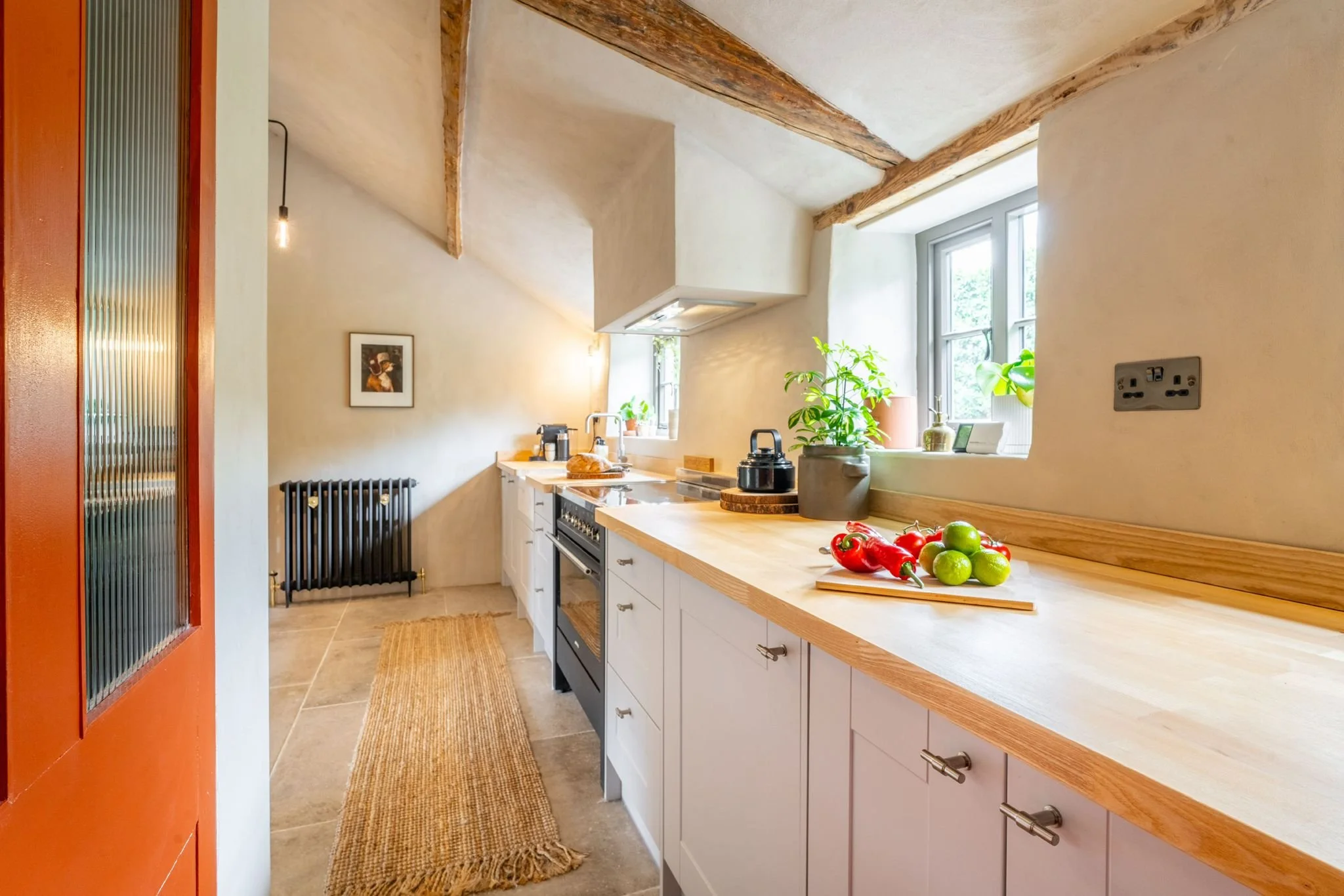 A cozy kitchen with sloped ceilings, wooden beams, a white countertop, and a window letting in natural light. The countertop has a cutting board with red peppers and green limes, and potted plants on the windowsill. There is an artwork on the wall an