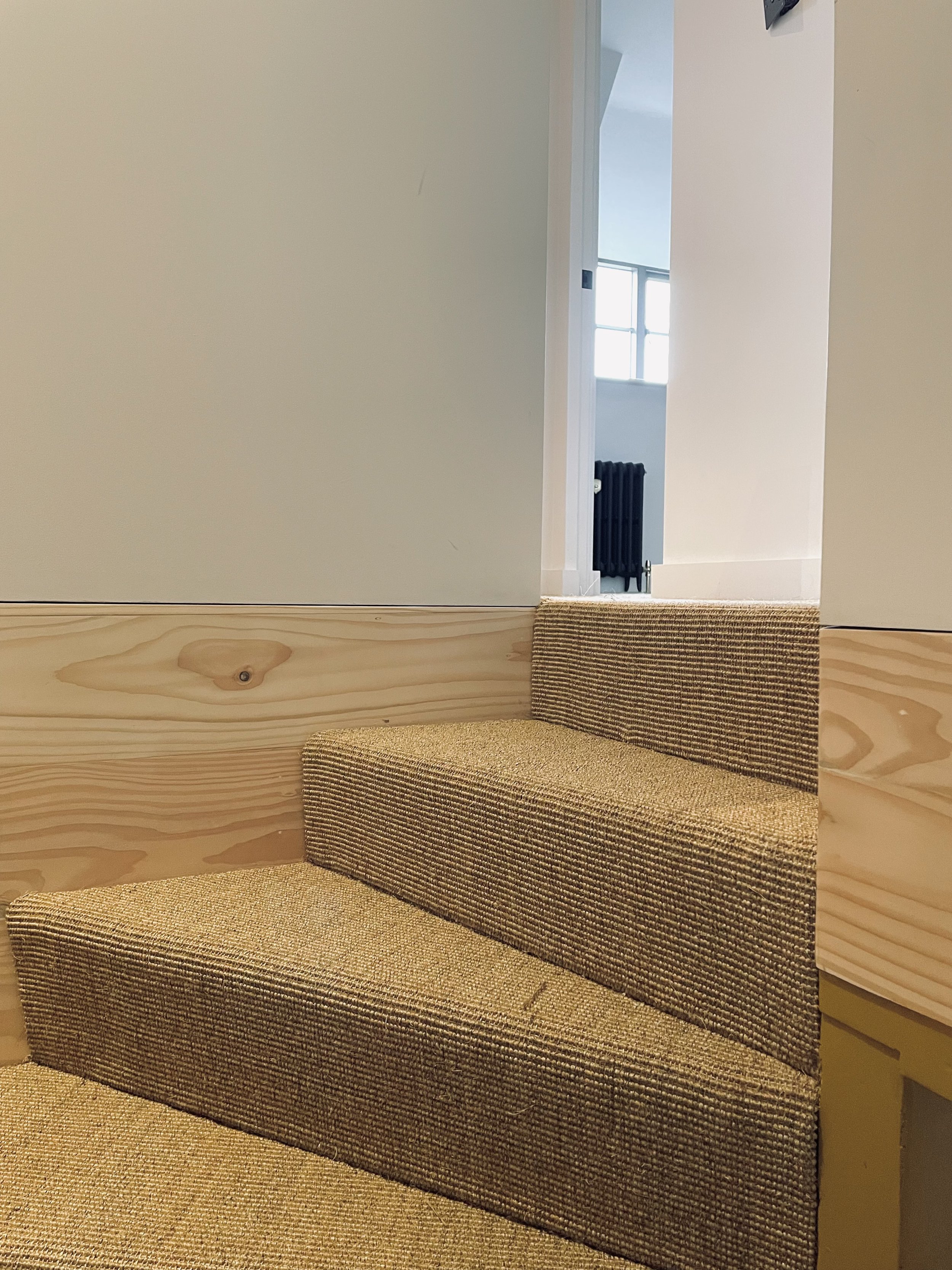 A staircase with a brown textured carpet, greenish wooden sides, and a beige wall, leading to a doorway that opens into a room with a window and a black radiator.