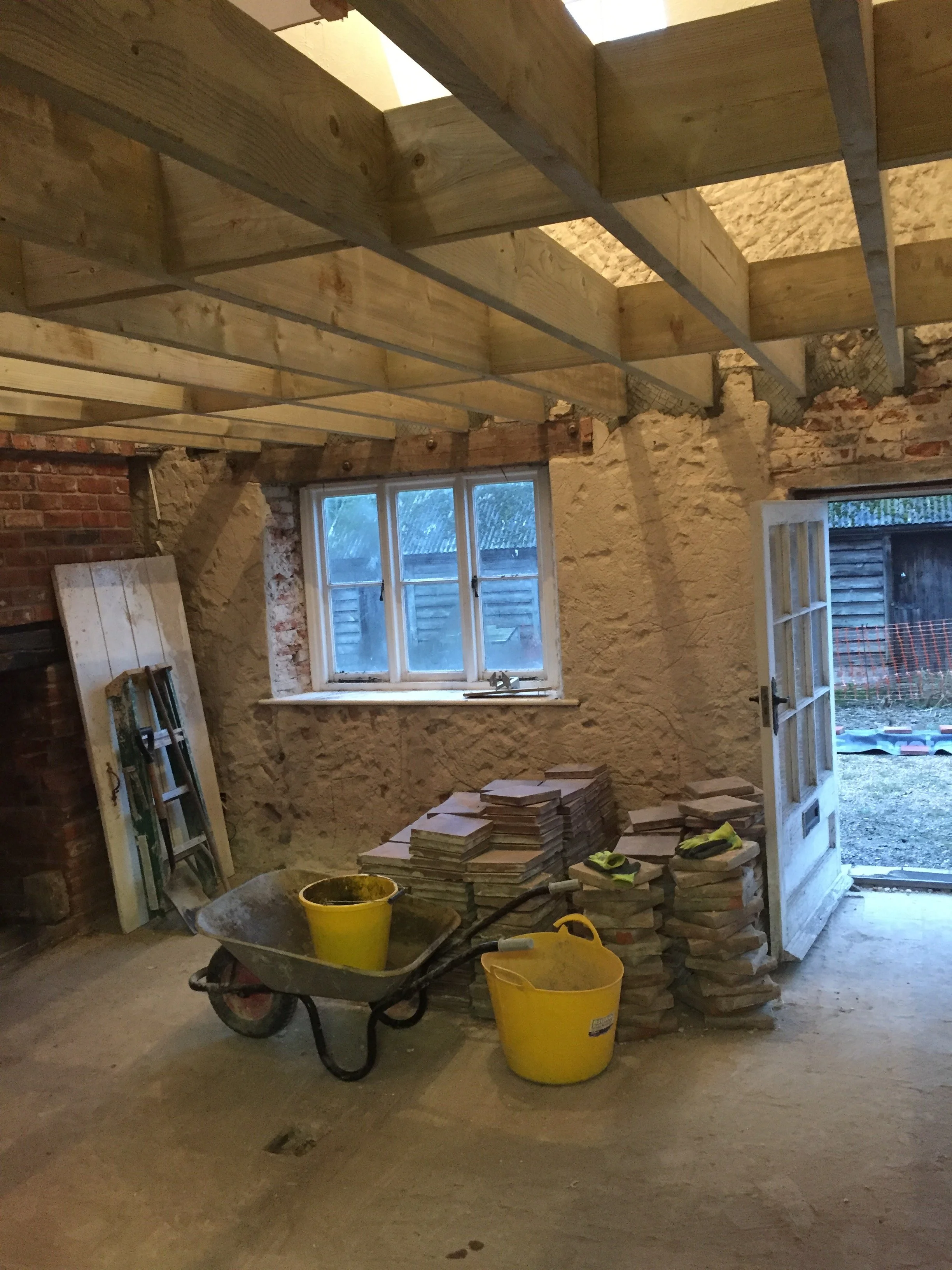 Interior of a room under renovation with exposed brick and stone walls, a window, construction materials, and tools including a wheelbarrow and yellow buckets.