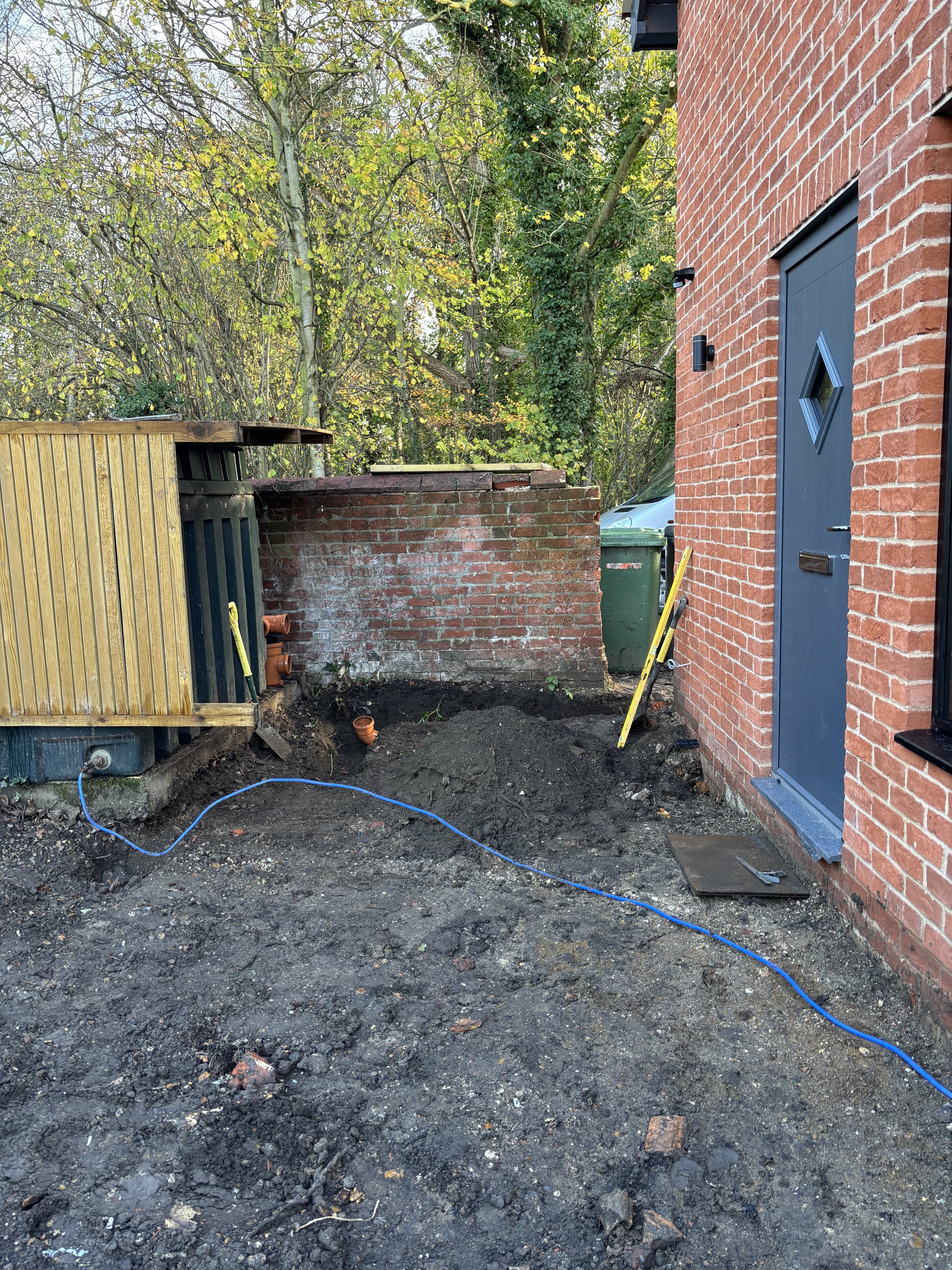 A backyard area undergoing construction with dark soil and exposed dirt, a brick wall of a house on the right, a green trash bin, a small wooden shed on the left, a tangled blue wire on the ground, and a backdrop of dense trees with yellow and green 