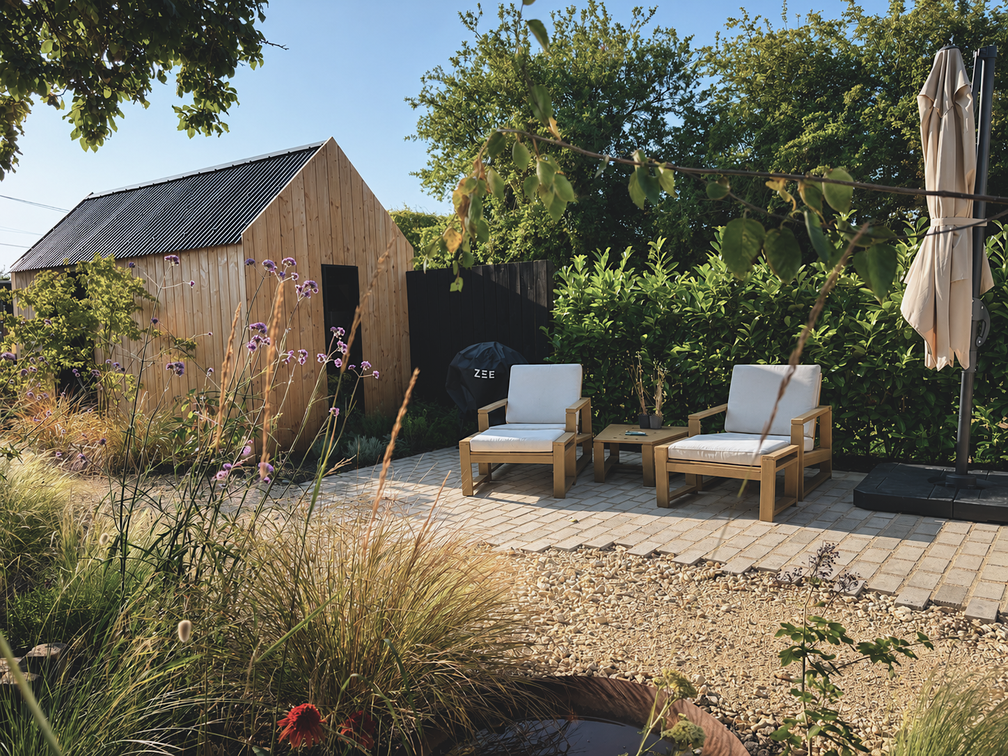 A backyard patio with two wooden chairs with white cushions, a small table between them, a barbecue grill labeled 'ZEE,' and a closed umbrella in a sunny garden with green bushes, trees, and a small wooden shed.