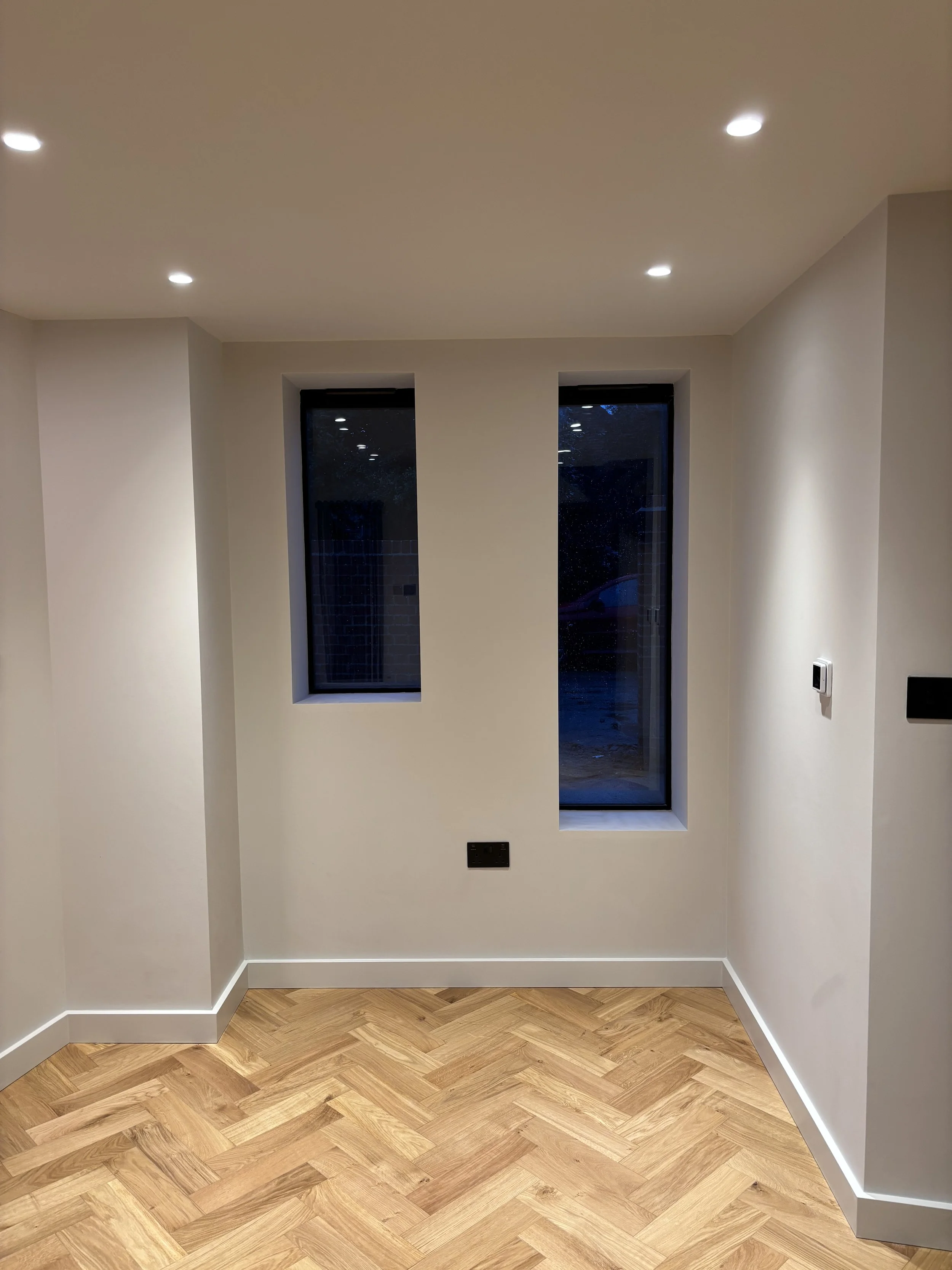 Modern interior room with white walls, two tall narrow windows, hardwood herringbone floor, recessed ceiling lights, and a thermostat on the wall.