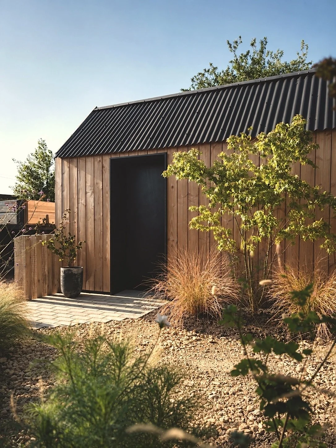 A small wooden shed with a black metal roof, surrounded by plants and a gravel yard.