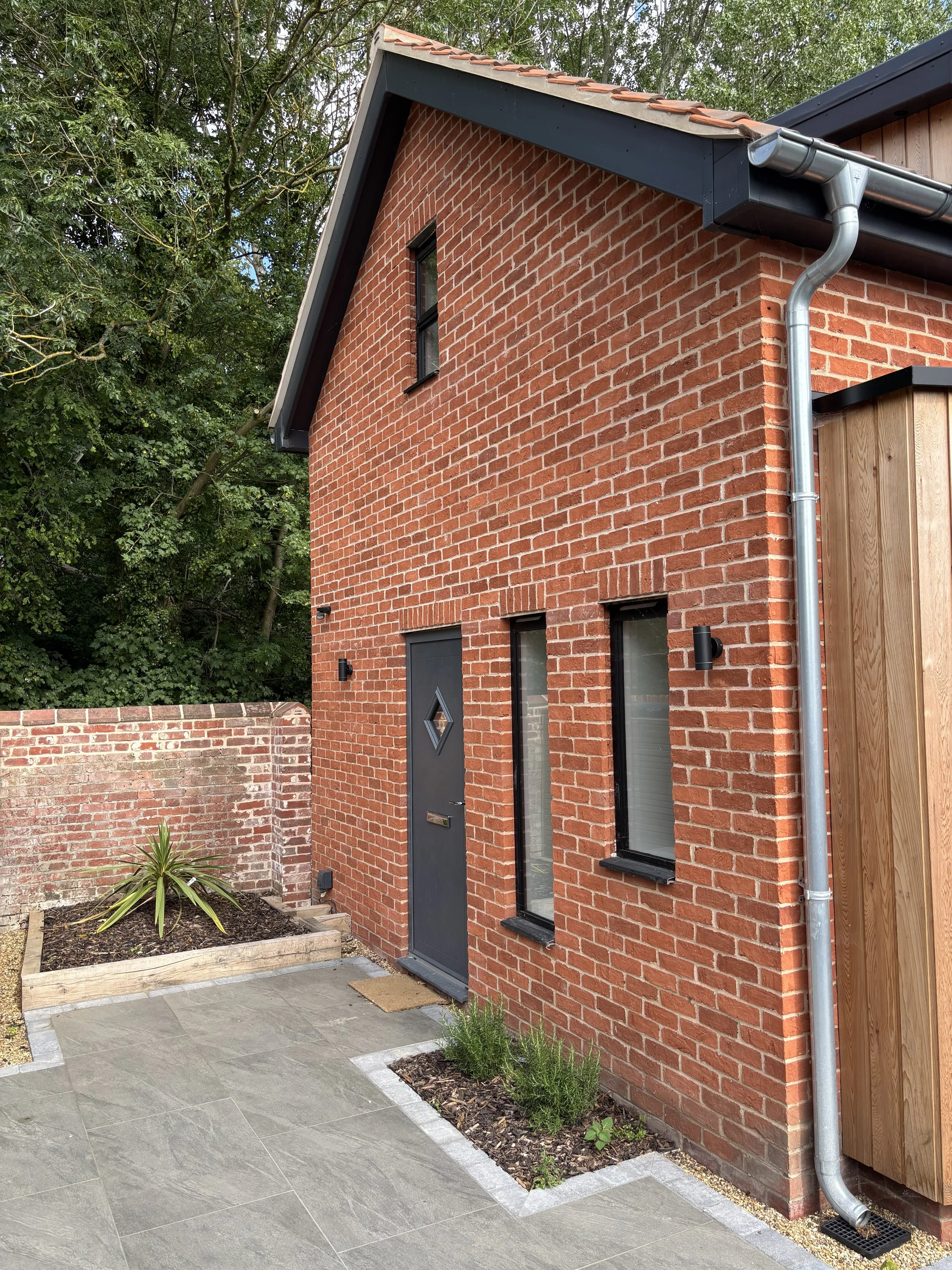 Modern brick house with black door and window frames, metal rain gutter, small garden bed with plants, stone patio, and trees in the background.