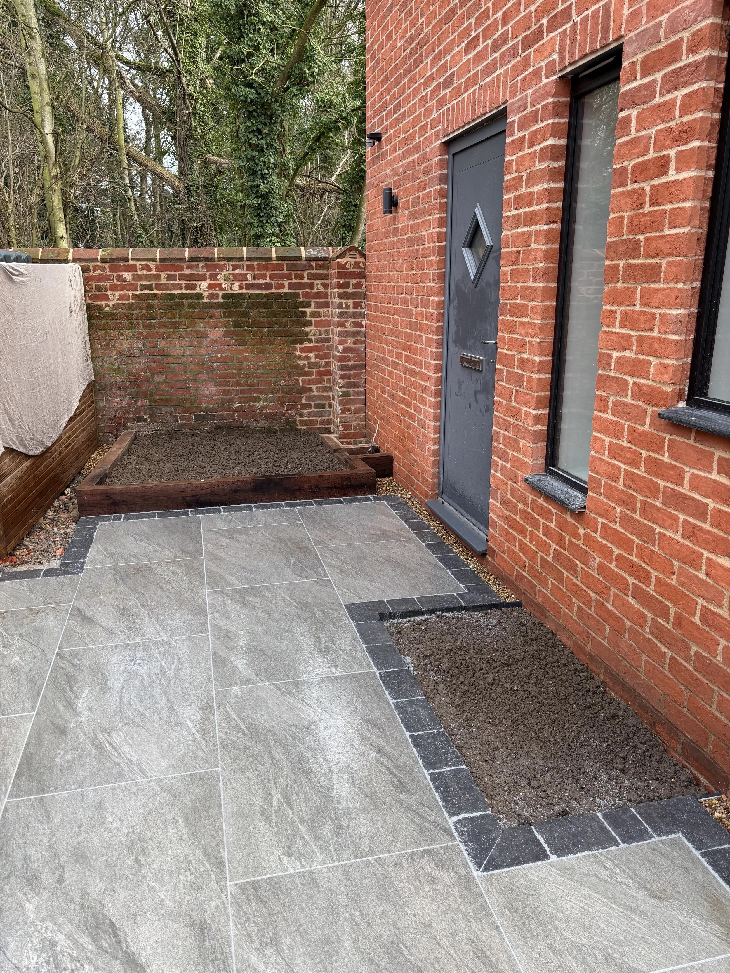 A patio with large gray tiles bordered by smaller dark gray tiles, adjacent to a brick house with a dark gray door and window, and a small garden bed with brown soil.