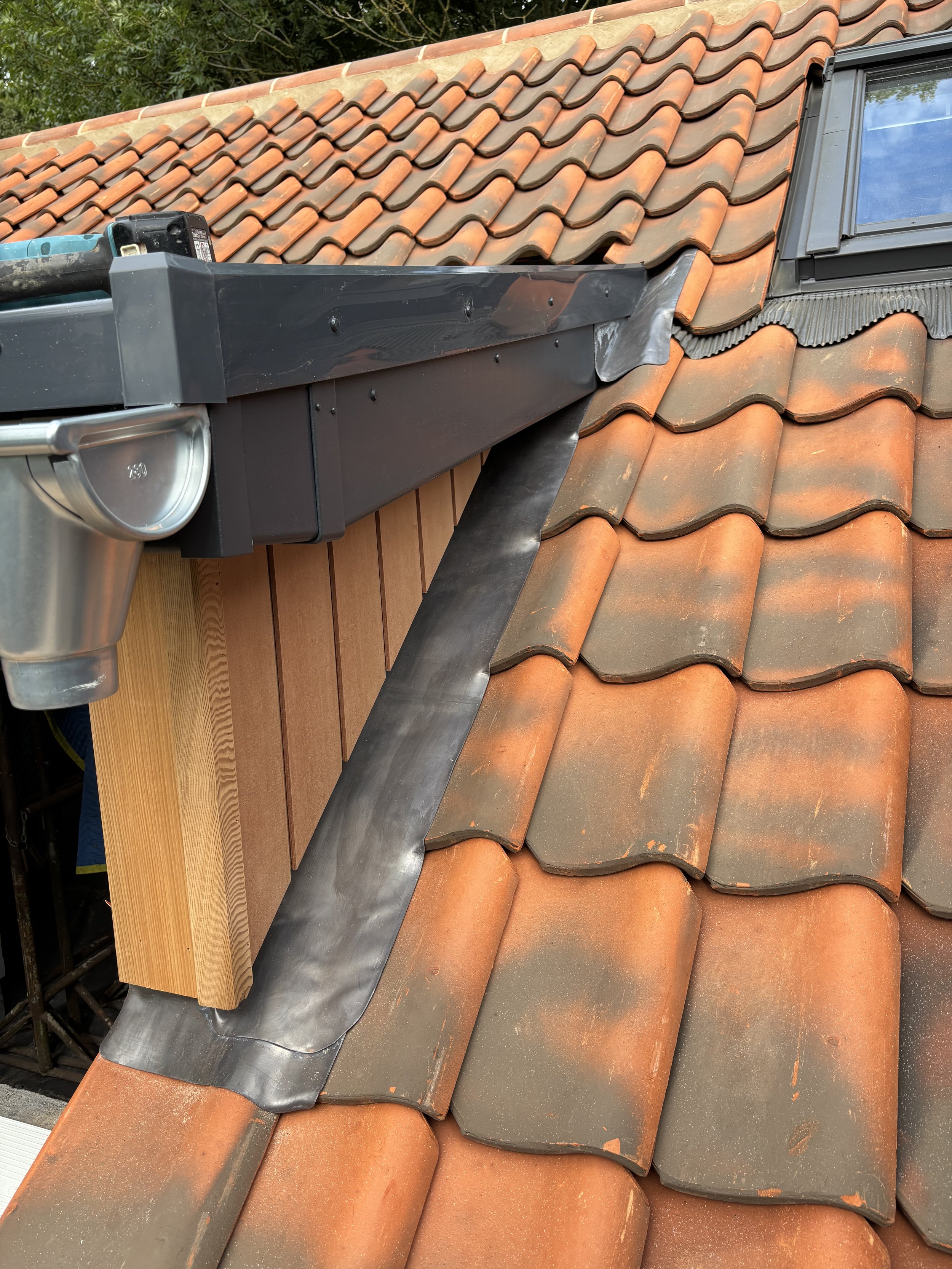 A section of a roof with orange clay tiles, a black metal rain gutter, and a dormer window with a dark frame and blue glass.