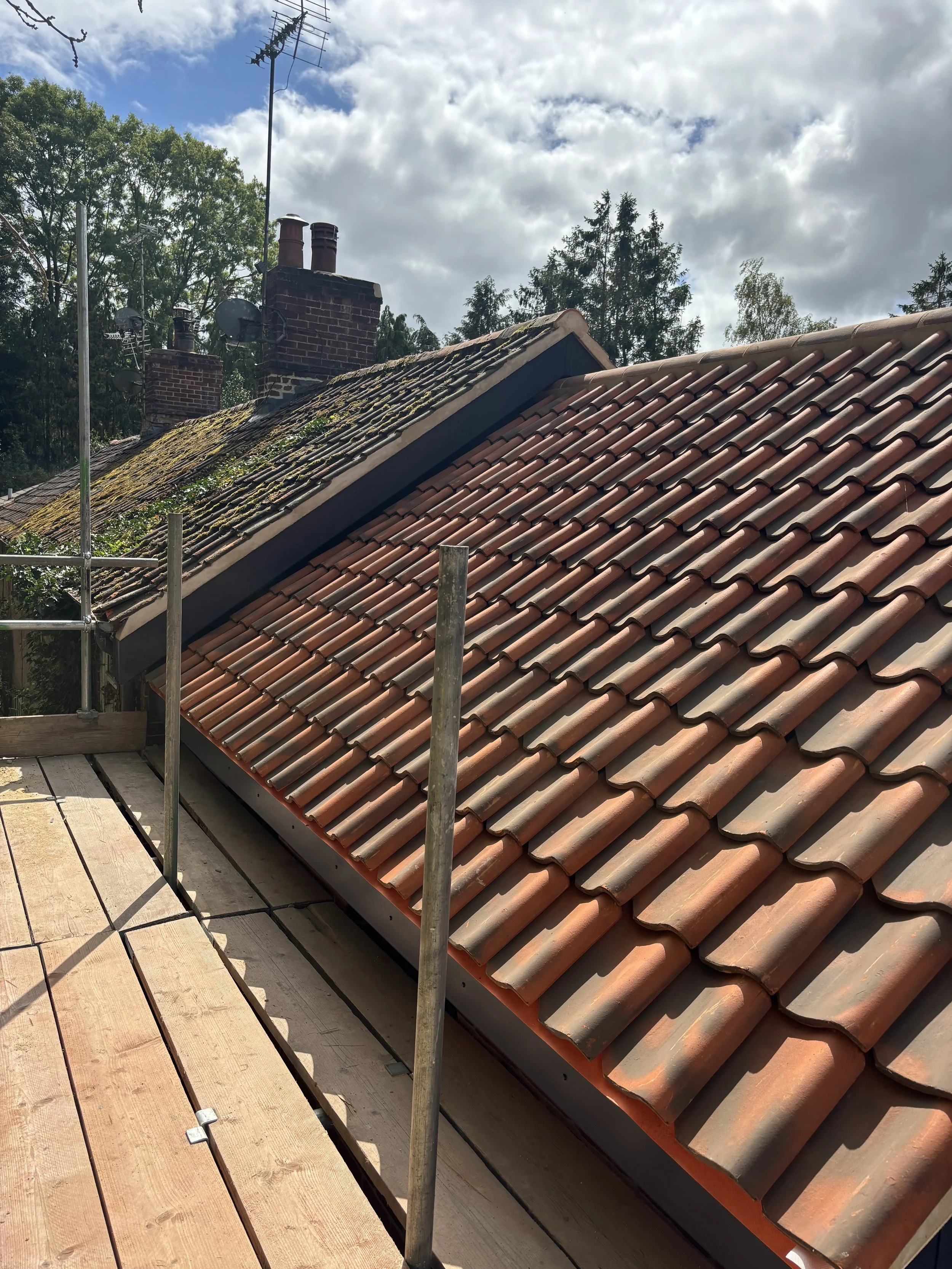 Rooftop with new tiled roof in progress, scaffolding, and chimney stacks with TV antennas against cloudy sky and trees.