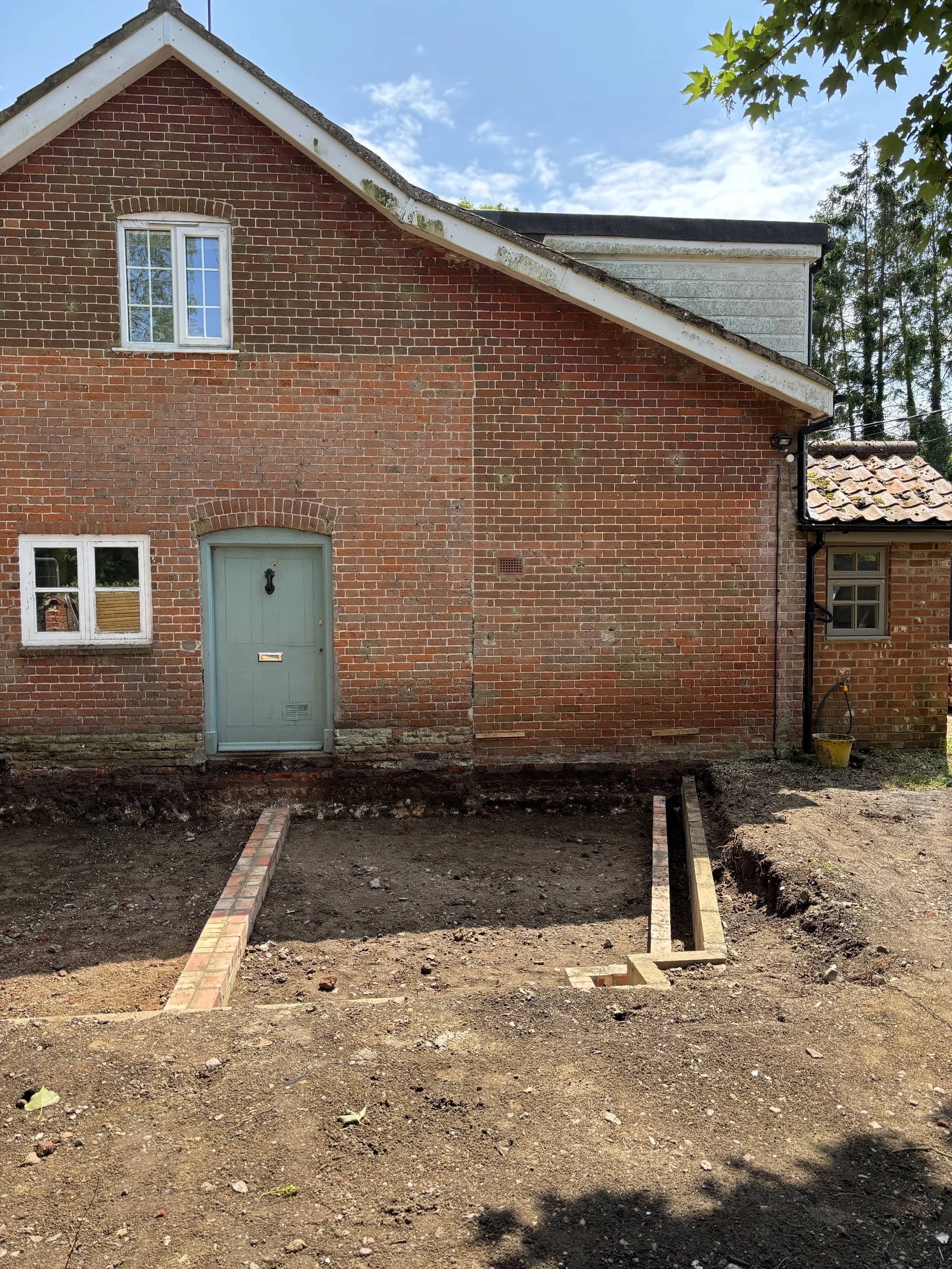 Backyard of a brick house under renovation, with an empty dugout lawn area, brick border edge, and some construction materials and tools visible at the edges.