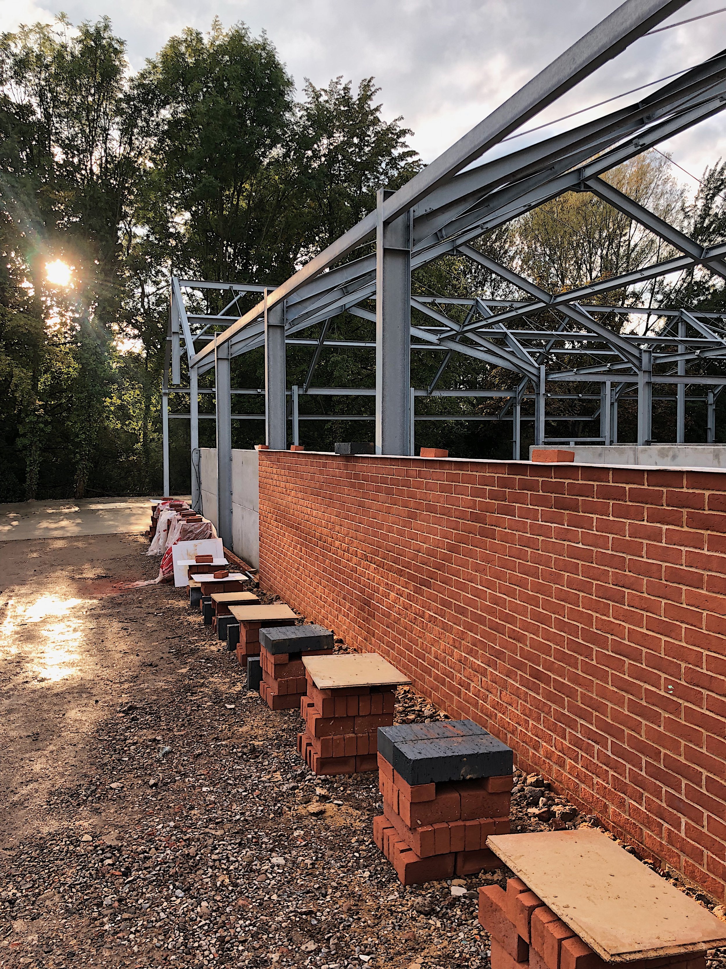 Construction site with a brick wall under construction, steel framework for a building's roof, and bricks stacked along the wall, with trees and a cloudy sky in the background.