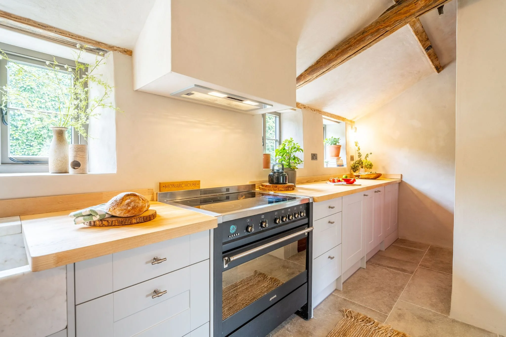 Kitchen with wooden countertops, white cabinets, and a black oven. Sunlight enters through windows with greenery outside. Decor includes potted plants and a loaf of bread on a cutting board.