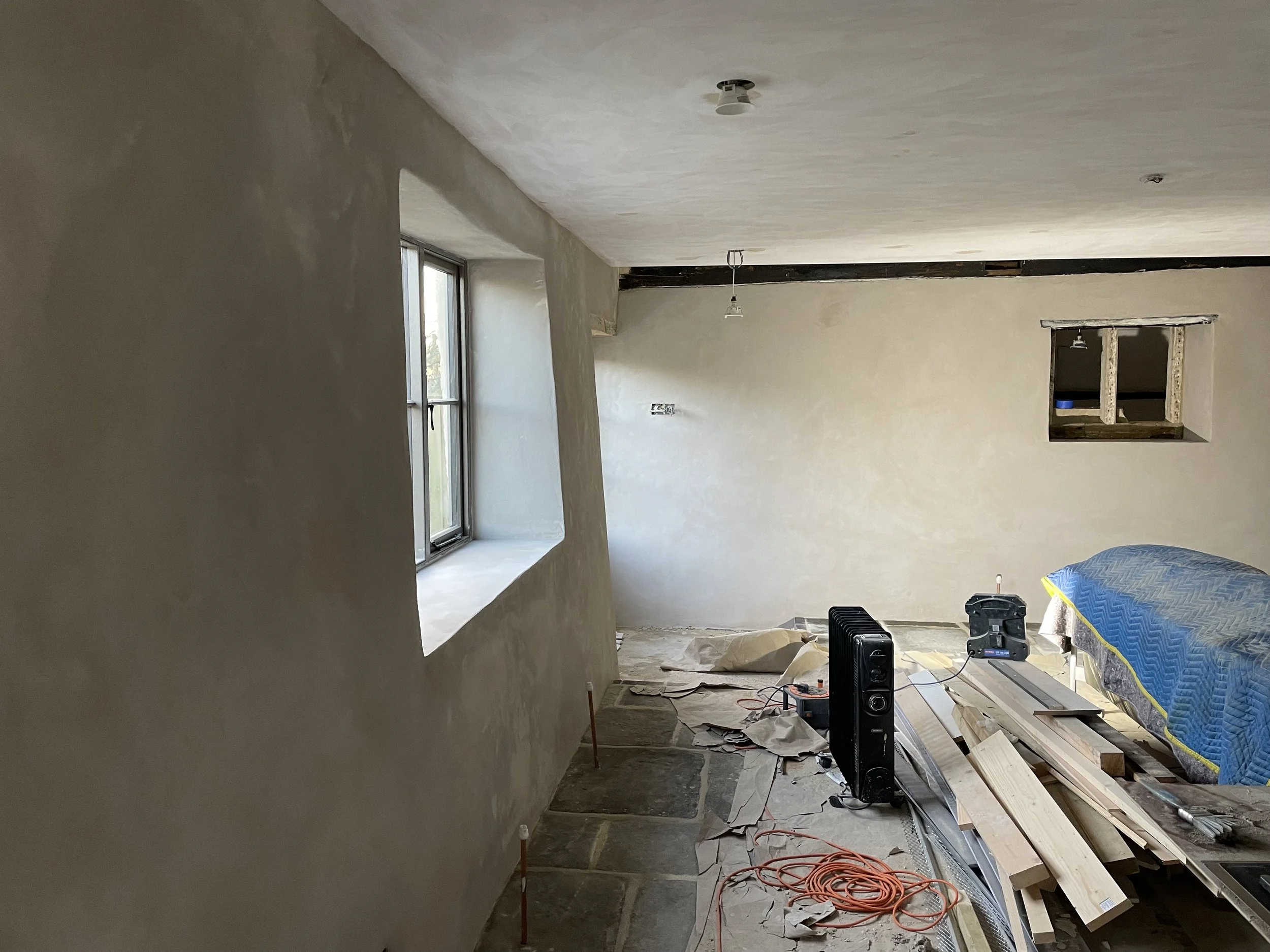 Interior of a house under renovation with unfinished walls, a window, construction materials, and tools scattered on the floor.