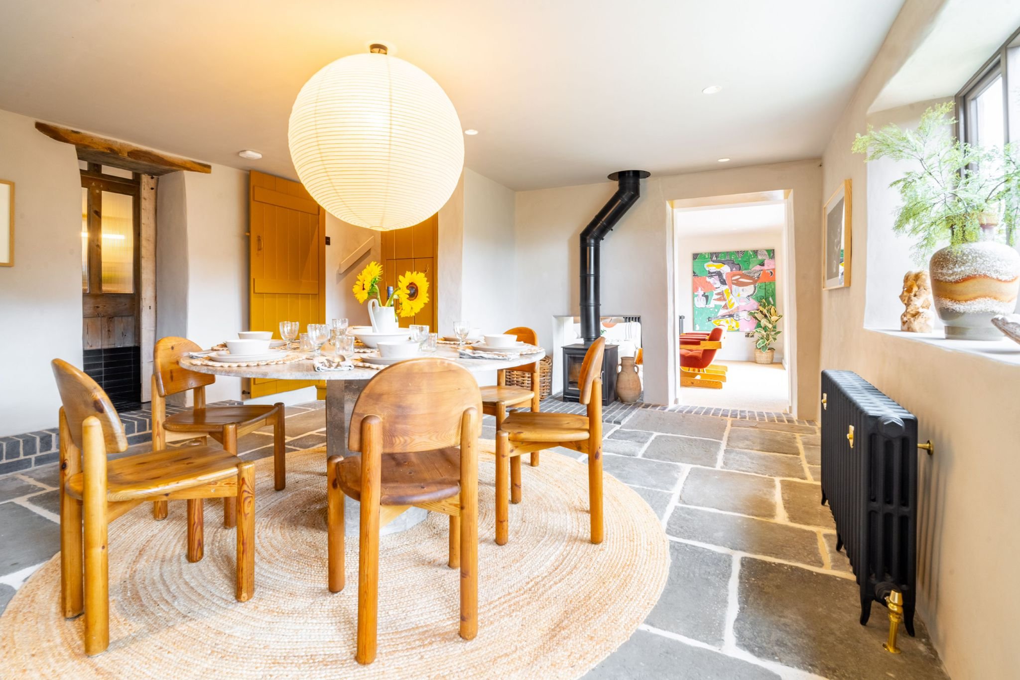 Dining area with a round table set with white bowls, glasses, and a white pitcher with sunflowers. Wooden chairs surround the table, illuminated by a large paper lantern pendant light. The room has a stone floor, a wood stove pipe, and decorative pot
