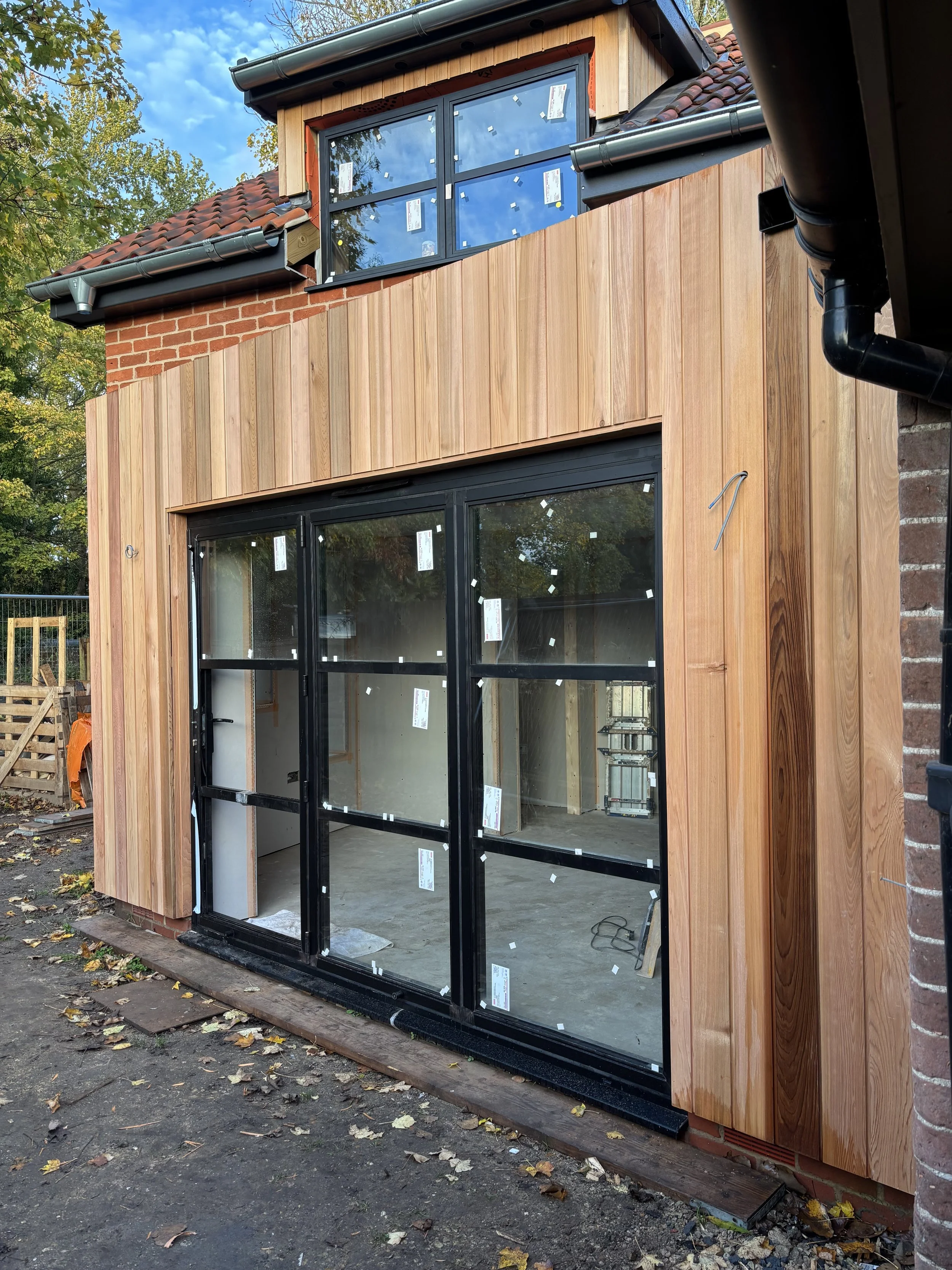 Front view of a house under construction with wooden siding, black window frames, and partially installed roof with red tiles. Leaves and construction materials are on the ground.