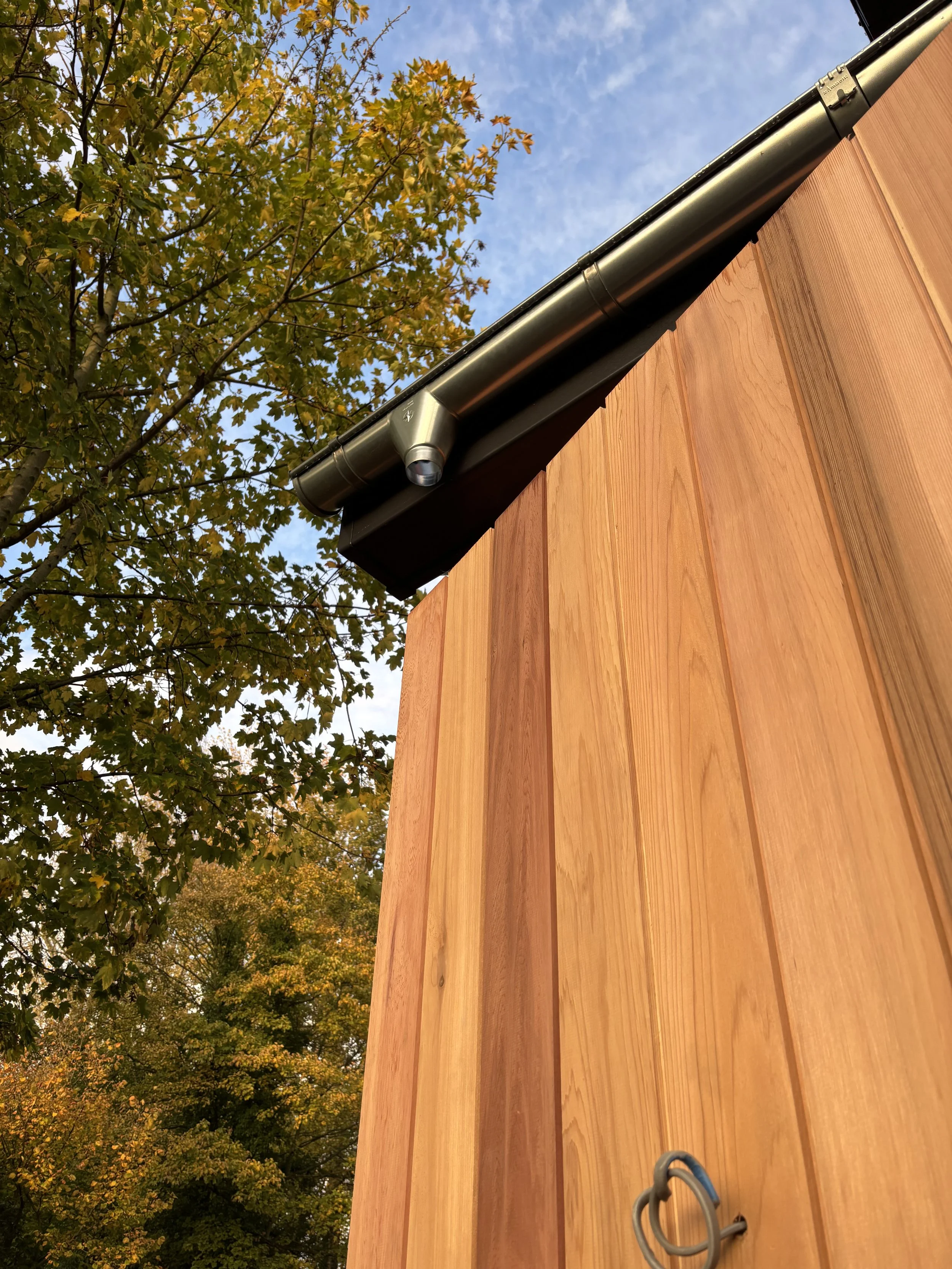 Close-up of the corner of a wooden building with a metal rain gutter, with green trees and a blue sky with clouds in the background.