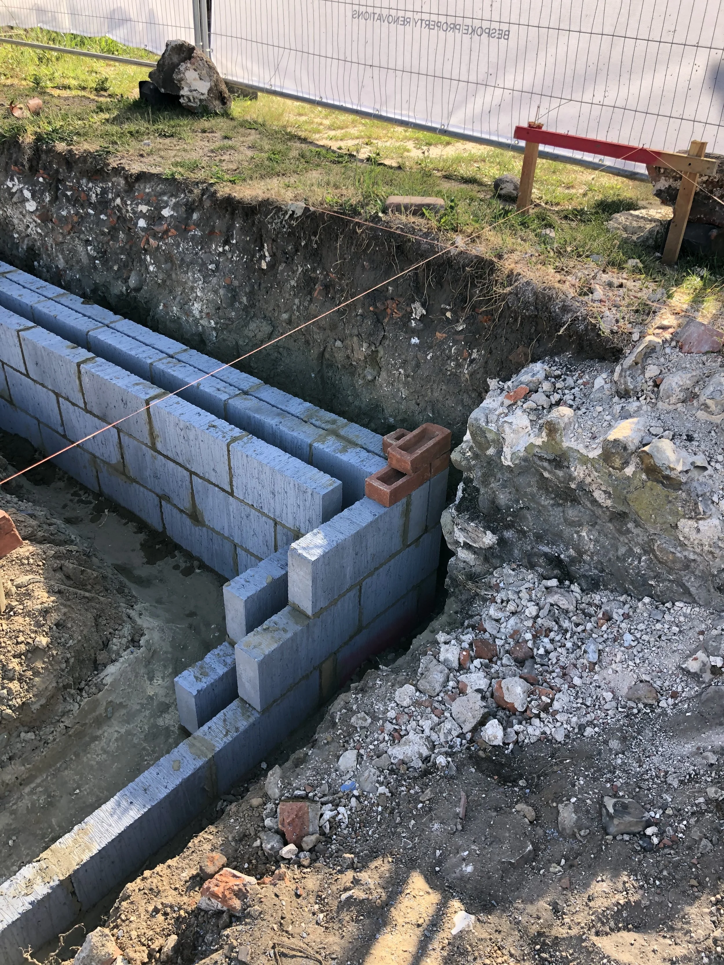 Construction site showing a concrete block wall under construction, with soil and rocks surrounding it, and a string line marking the layout.