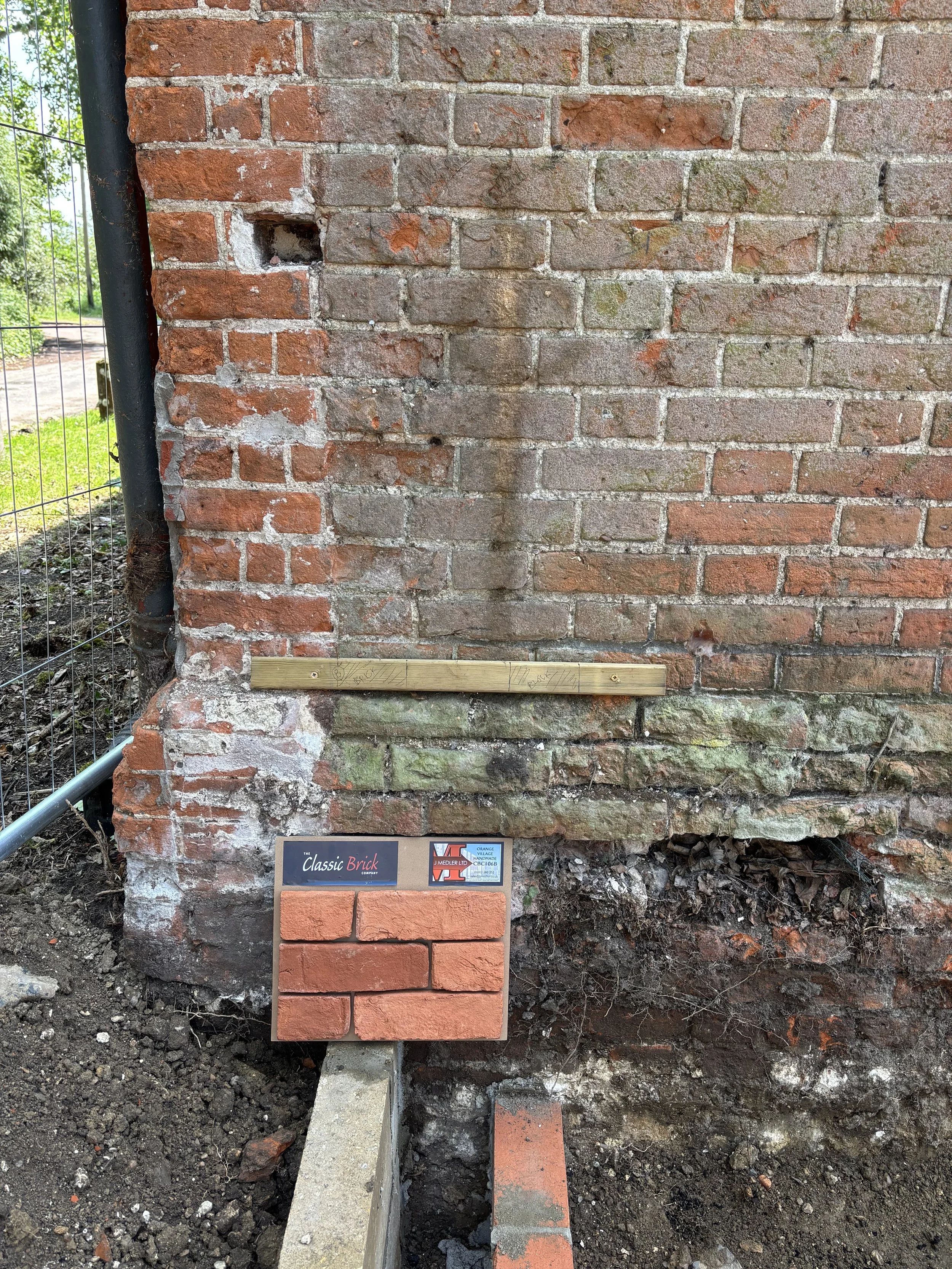 Close-up view of a brick wall under construction with a new section being built, showing a small brick wall with a plaque that reads 'The Classic Brick Company.' There are some construction tools and materials visible, and the ground is dug up for th