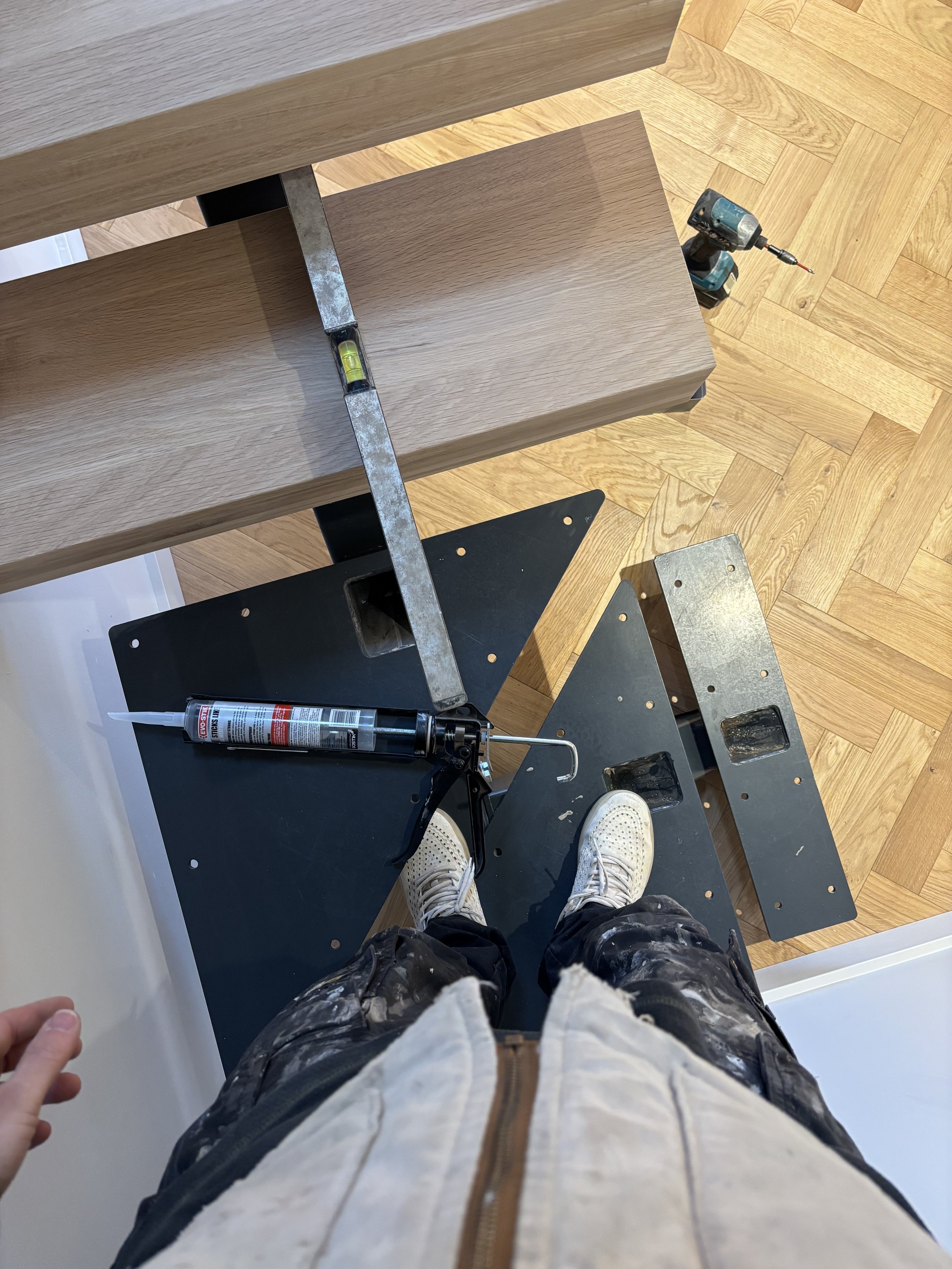 Overhead view of a person standing on a black scaffolding with their feet visible, wearing white sneakers, surrounded by tools and wooden flooring.