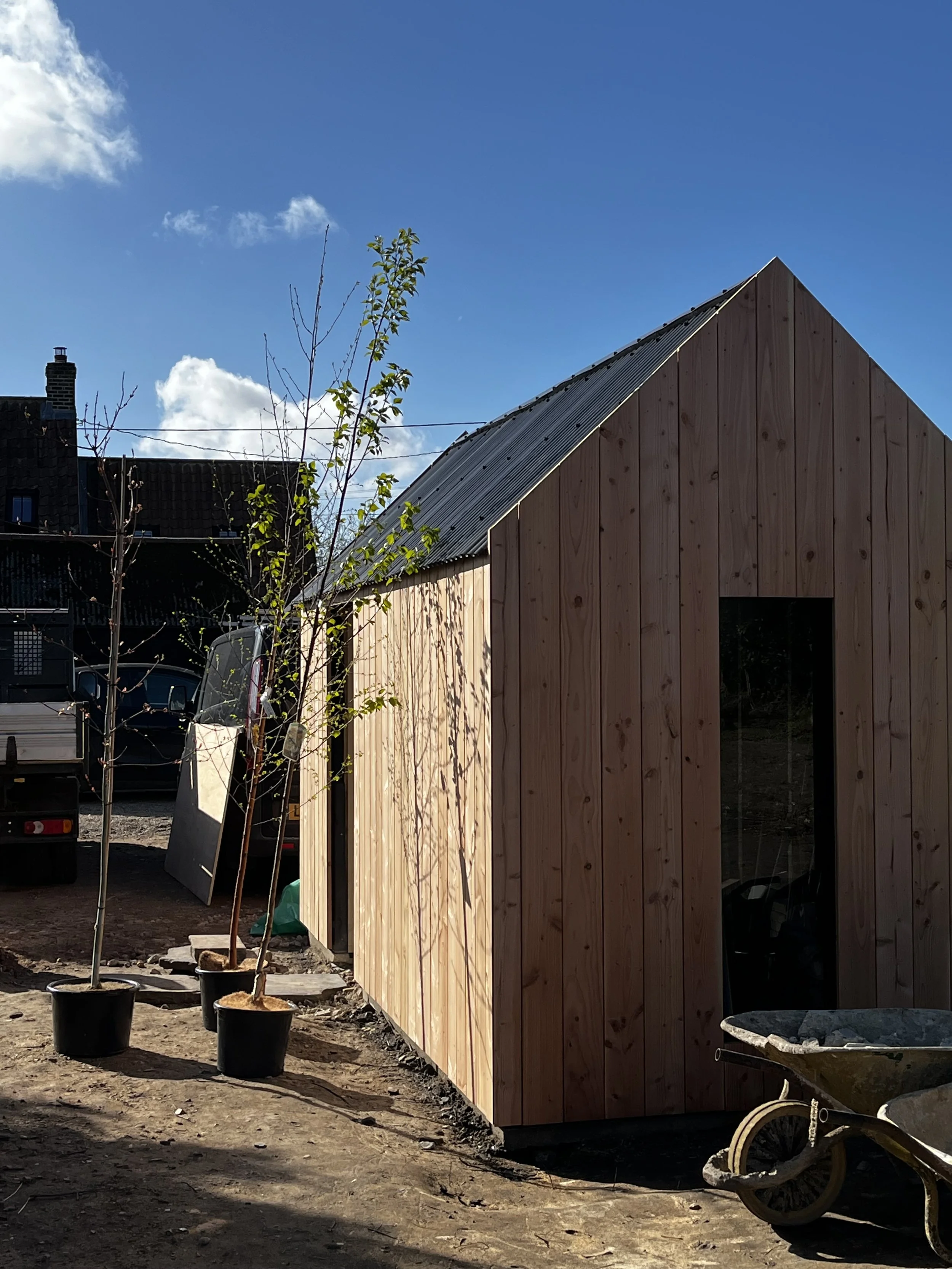 Construction site with a wooden building, potted young trees, a wheelbarrow, and a pickup truck under a blue sky with white clouds.