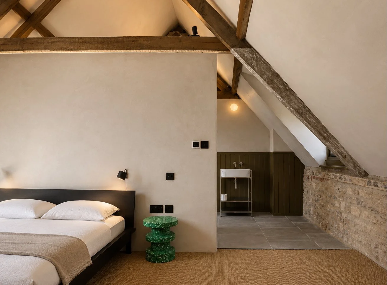 Modern bedroom with a bed, a black bedside lamp, and a green textured side table, with a rustic lofted ceiling and an open area with a small sink and exposed brick wall.
