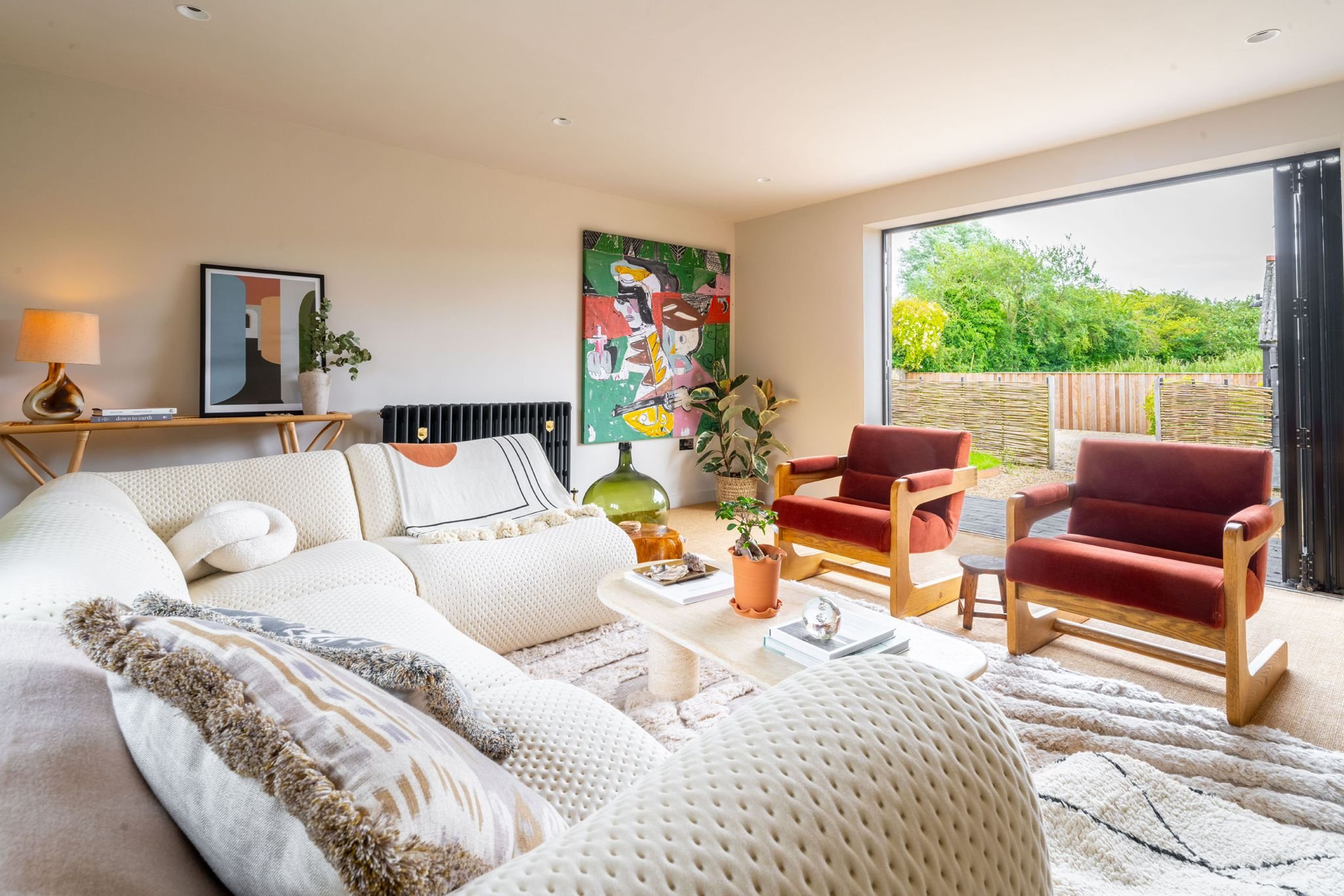 A cozy living room with a cream sofa, red armchairs, a coffee table, indoor plants, colorful artwork on the wall, and a large window with a view of a outdoor garden with greenery and a wooden fence.