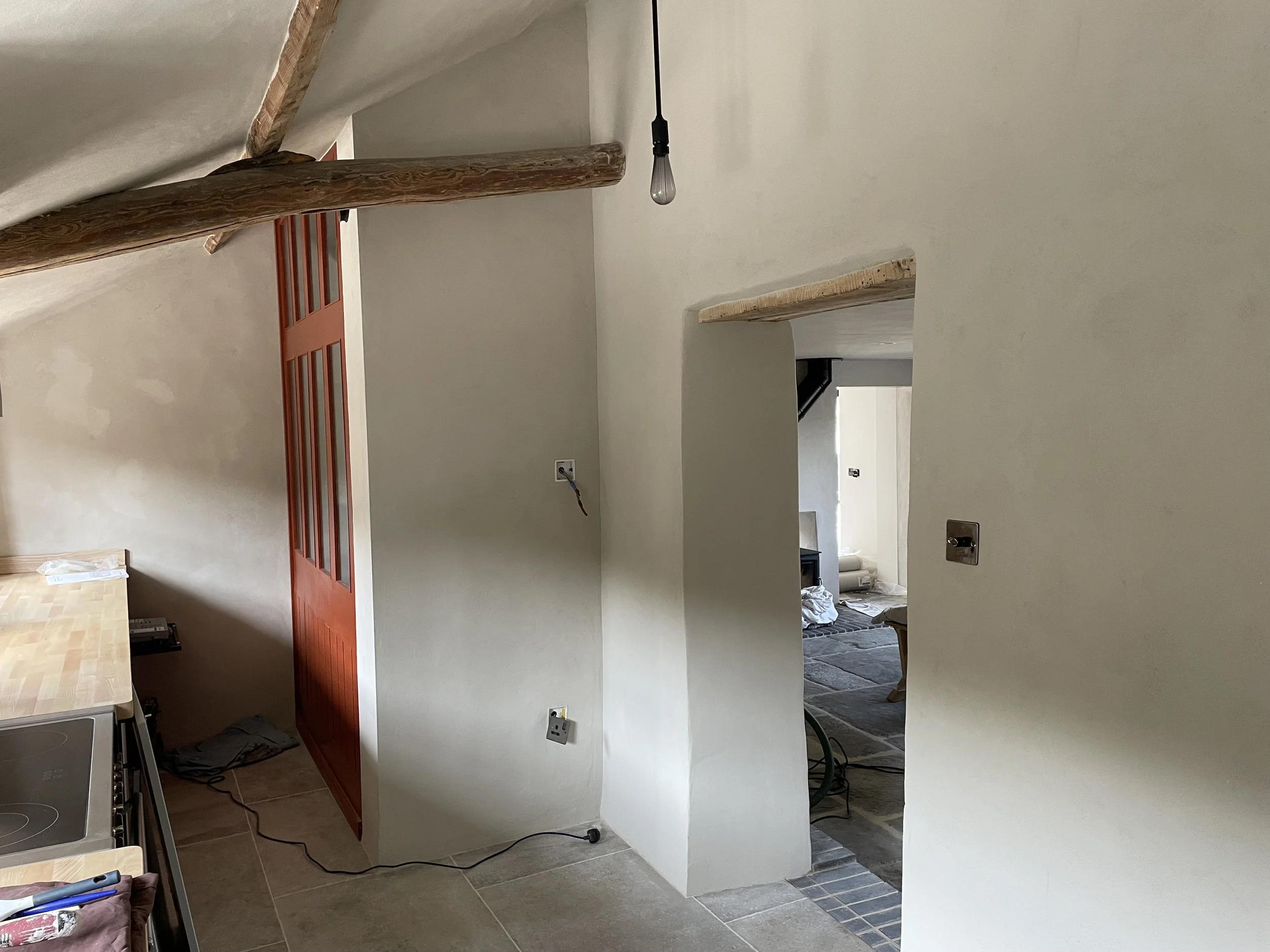 Interior view of a hallway with beige walls, a wooden ceiling beam, and a hanging light bulb. A red door is visible, and there is construction or renovation materials scattered around.