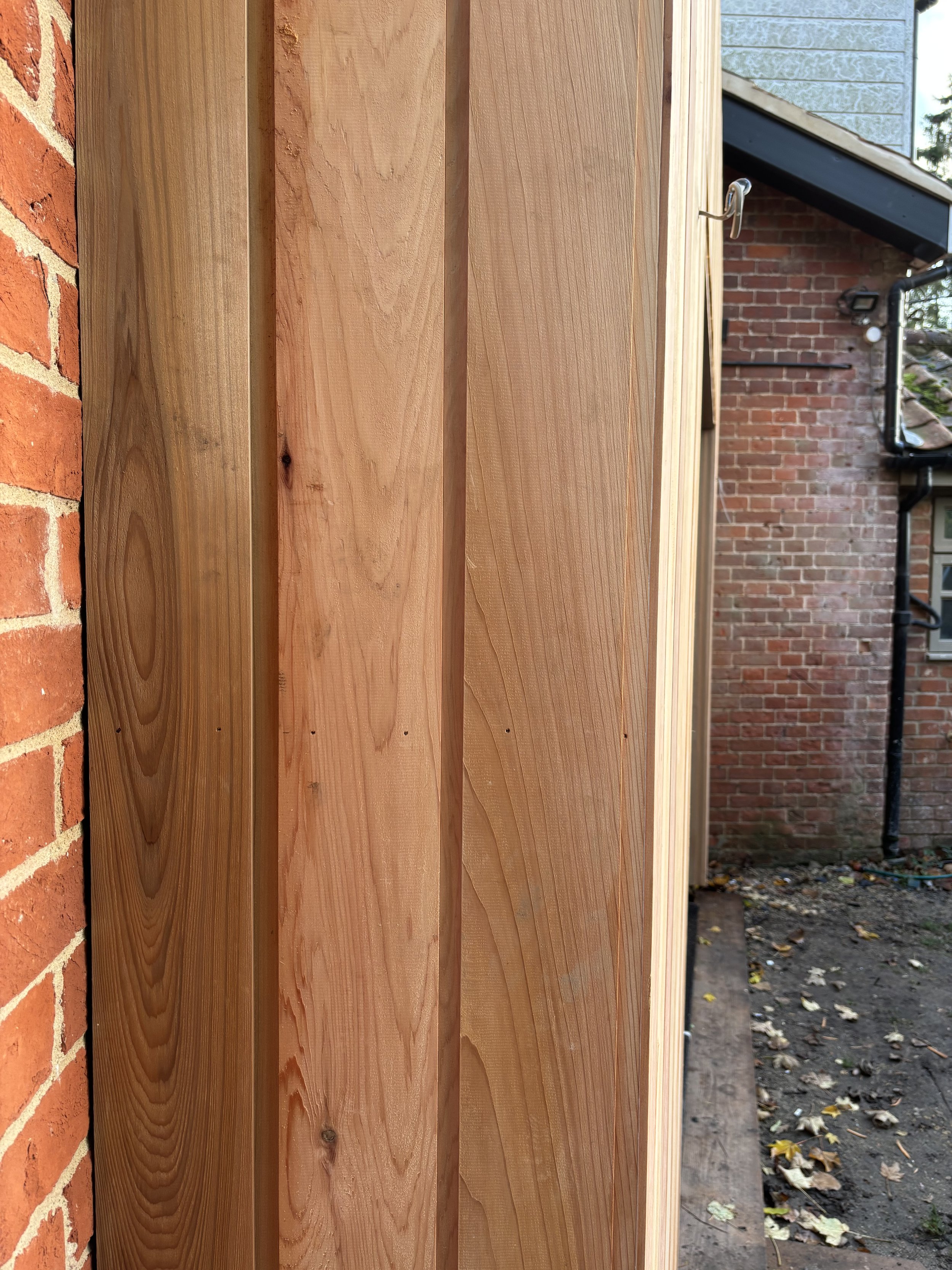 Close-up of vertical wooden planks attached to a brick wall, with a house and a drainpipe visible in the background.