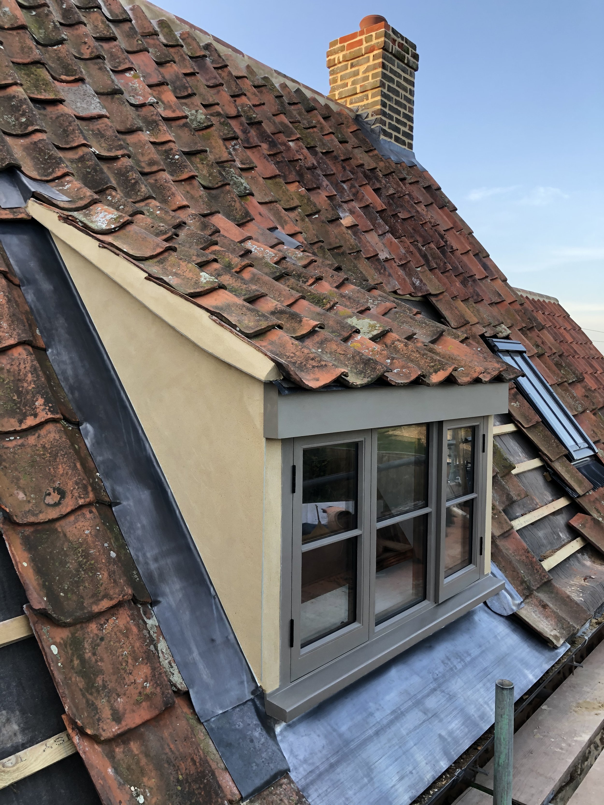 A rooftop with weathered, moss-covered red clay tiles. A newly installed window with a grey frame is protruding from the roof, surrounded by a partially completed roof renovation with some tiles missing. The sky in the background is clear and blue.