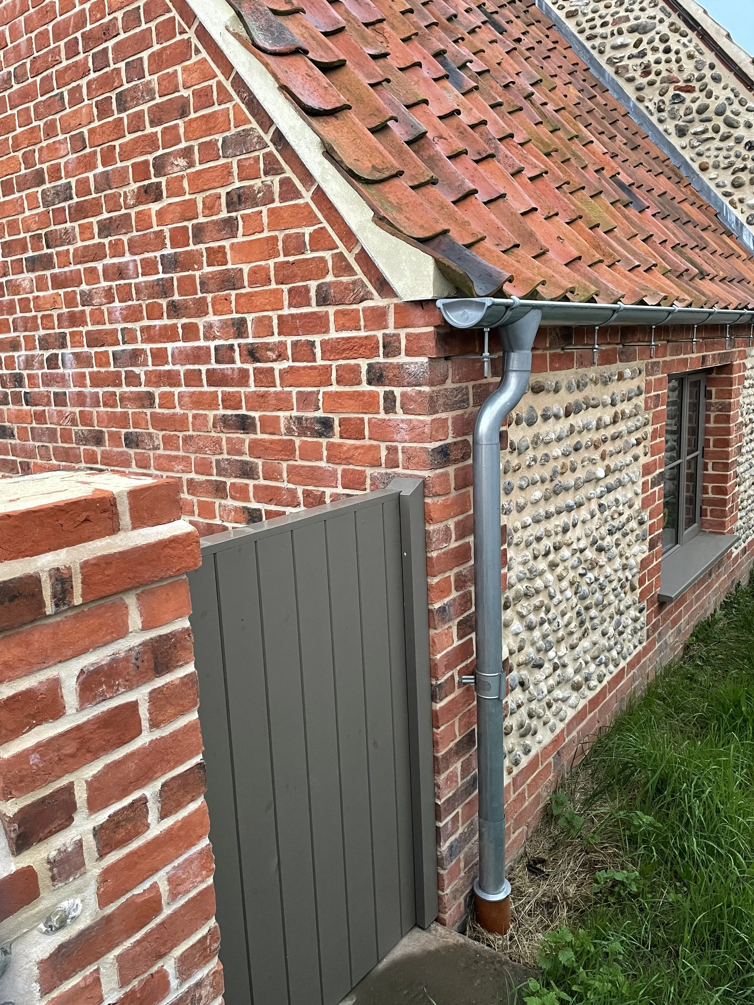 Close-up of a brick wall with a section of a roof, showing a rain gutter and downspout system, a small window, and a painted wooden door.