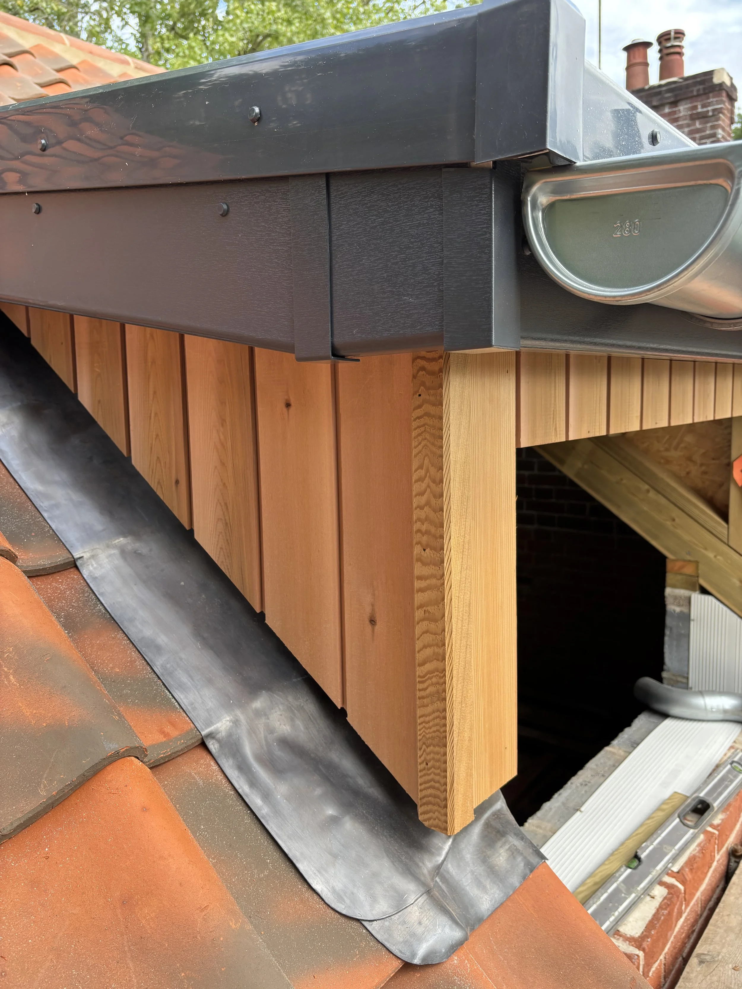 Close-up of a roof corner showing a black gutter, a section of wooden facade, and terracotta roof tiles, with chimney pipes and a tree in the background.