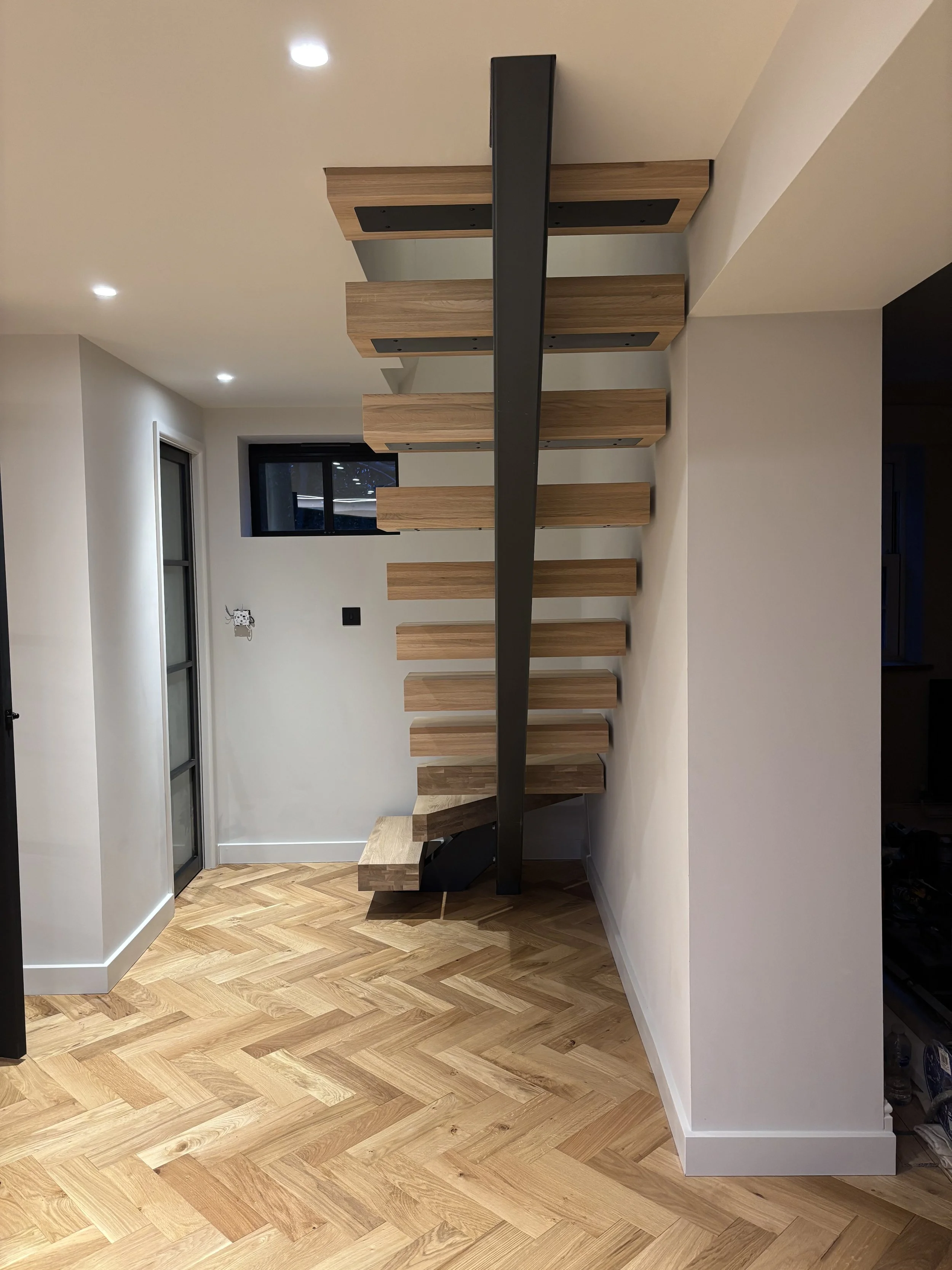 Modern staircase with wooden steps and a black support structure in a room with wooden flooring and white walls, overhead lighting, and a window.