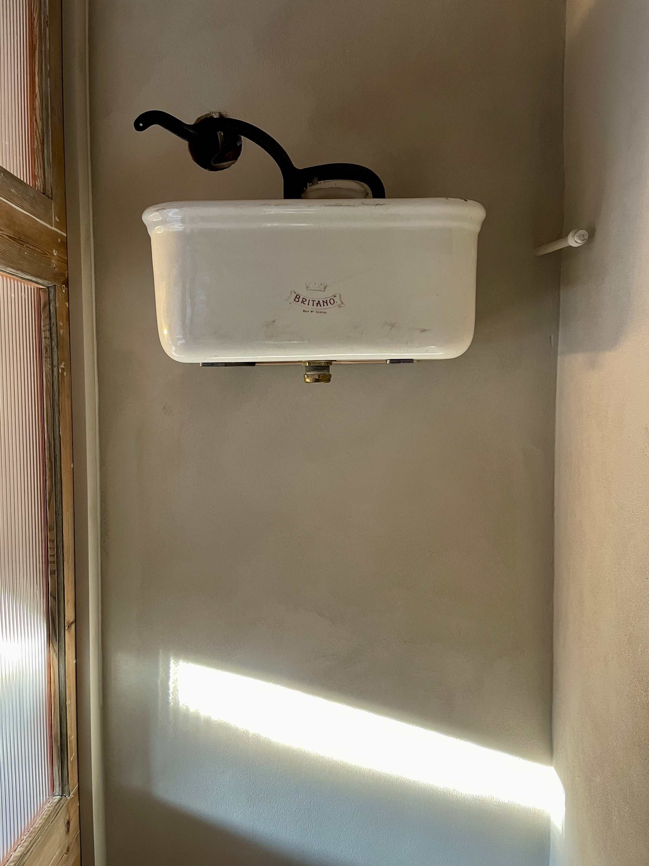 An old-fashioned white toilet tank mounted on a wall, with a black flush handle and a sticker that says 'Britano'.