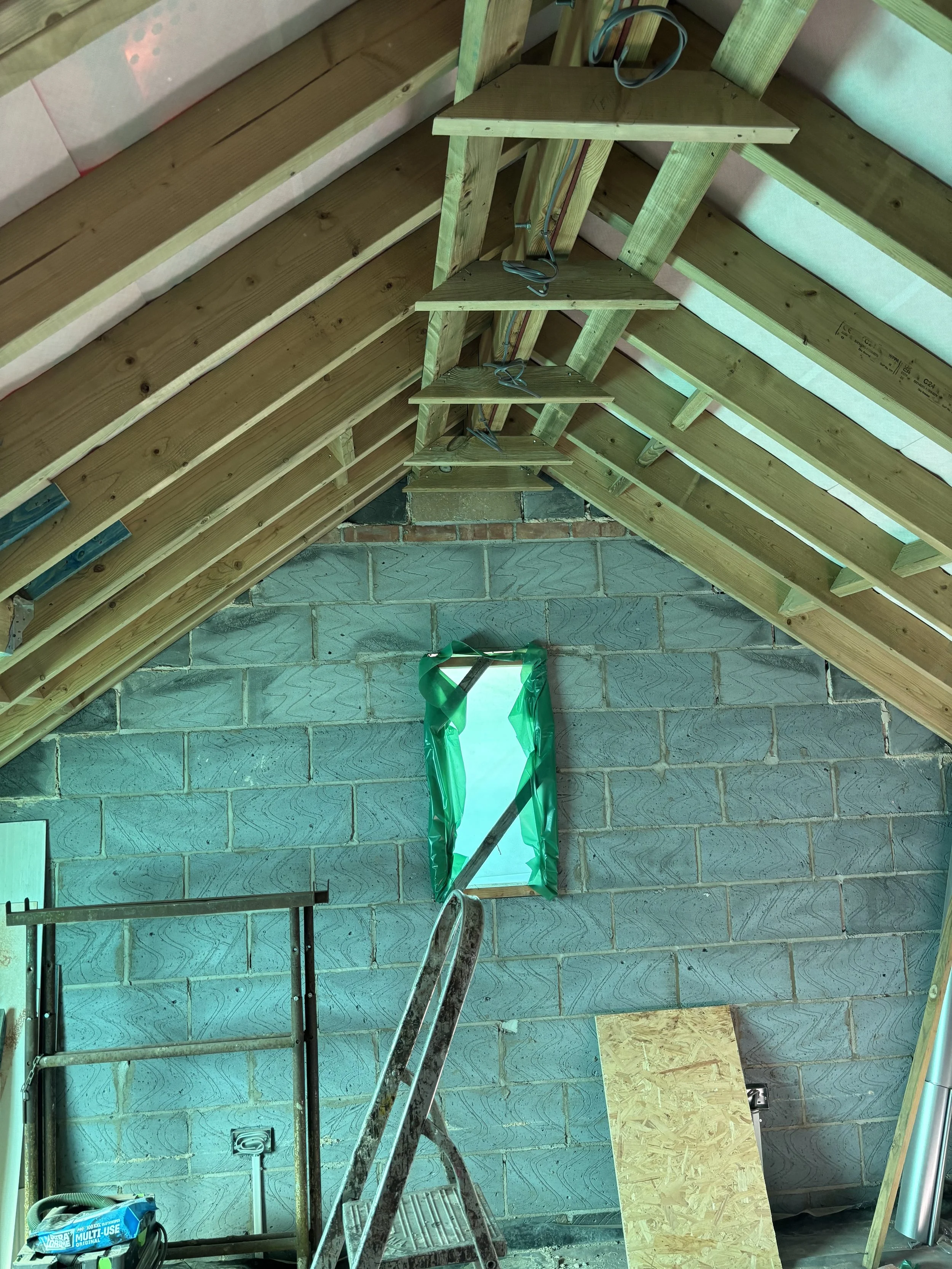 Attic space under construction with exposed wooden framing, a window covered partially with green plastic, a ladder, and construction tools.
