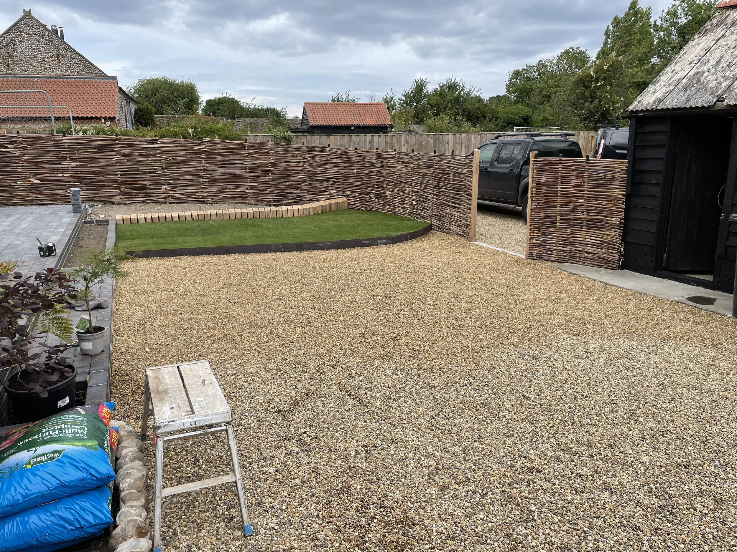 A backyard with a gravel ground, a small lawn area, and a woven wood privacy fence. There are garden plants with pots on the left, gardening supplies in the foreground, and a black shed on the right. A car is parked outside the fence, with houses and