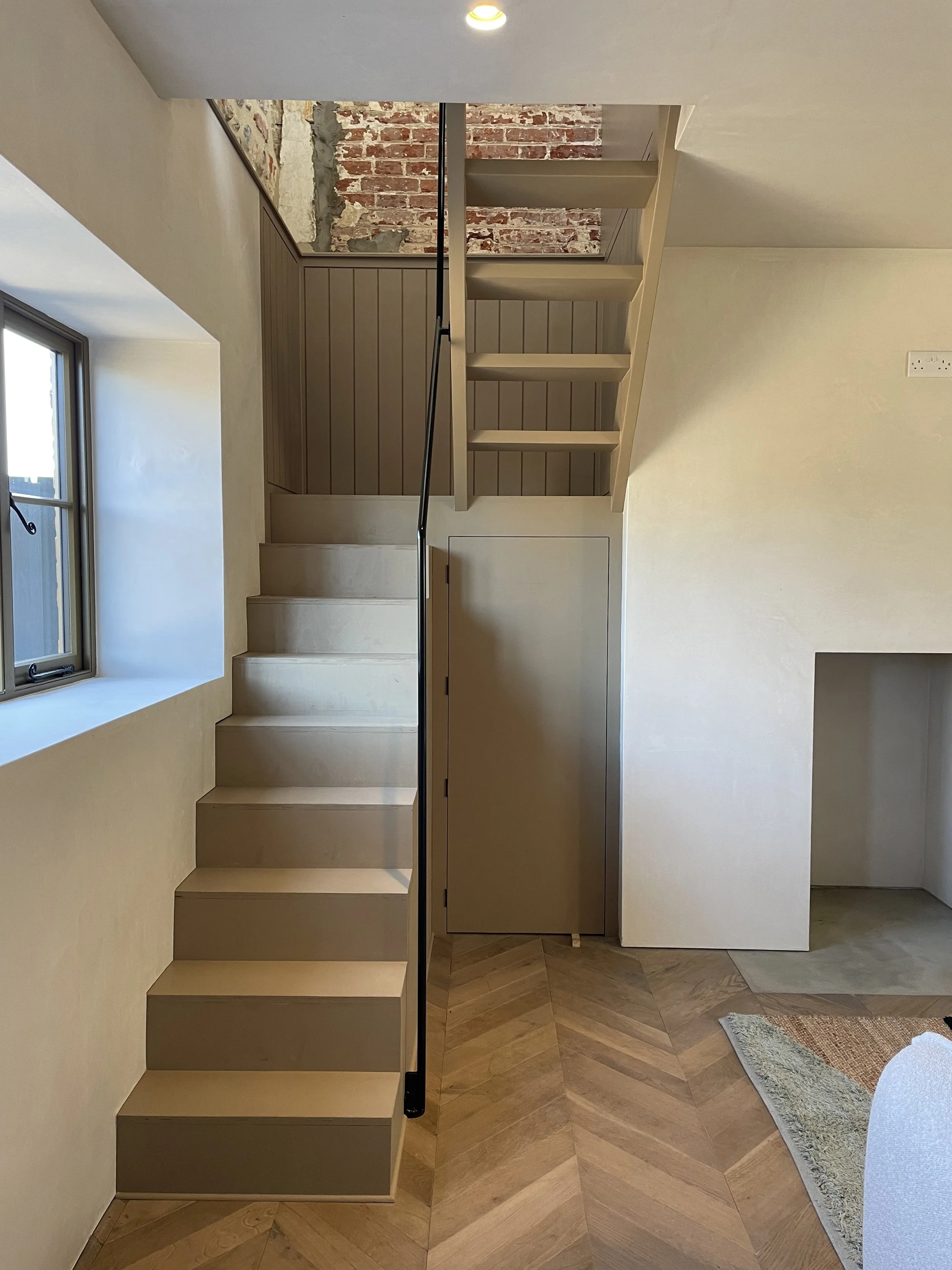 Interior view of a staircase with wooden steps, a black handrail, a window on the left, exposed brick wall at the top, and a small white door at the bottom of the stairs. A wooden floor and a rug are visible at the bottom right.
