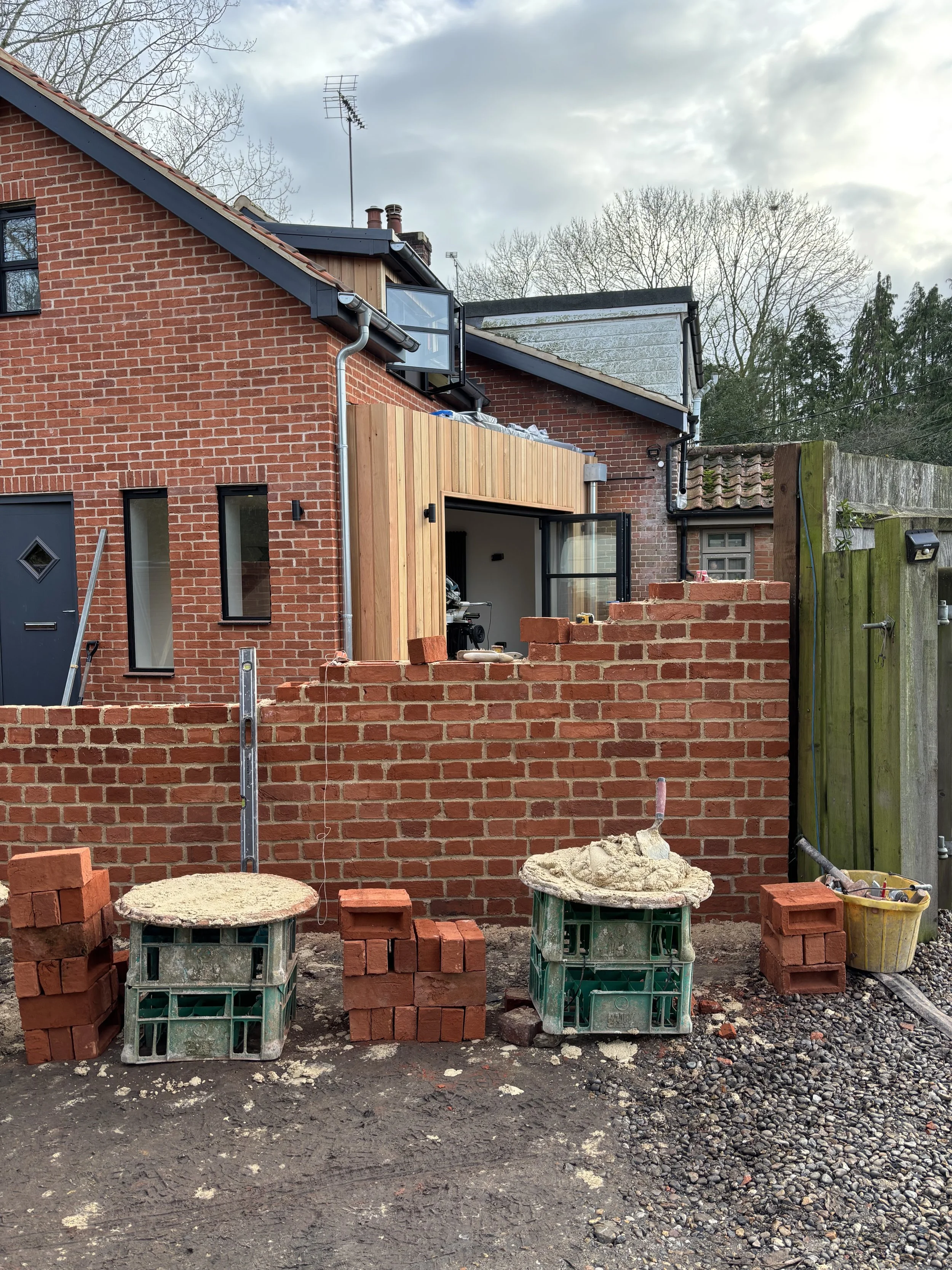 A construction site with a partially built red brick wall, stacks of bricks, and construction materials in front. The background shows a house with a brick exterior, windows, and a wooden extension, with trees and a cloudy sky above.