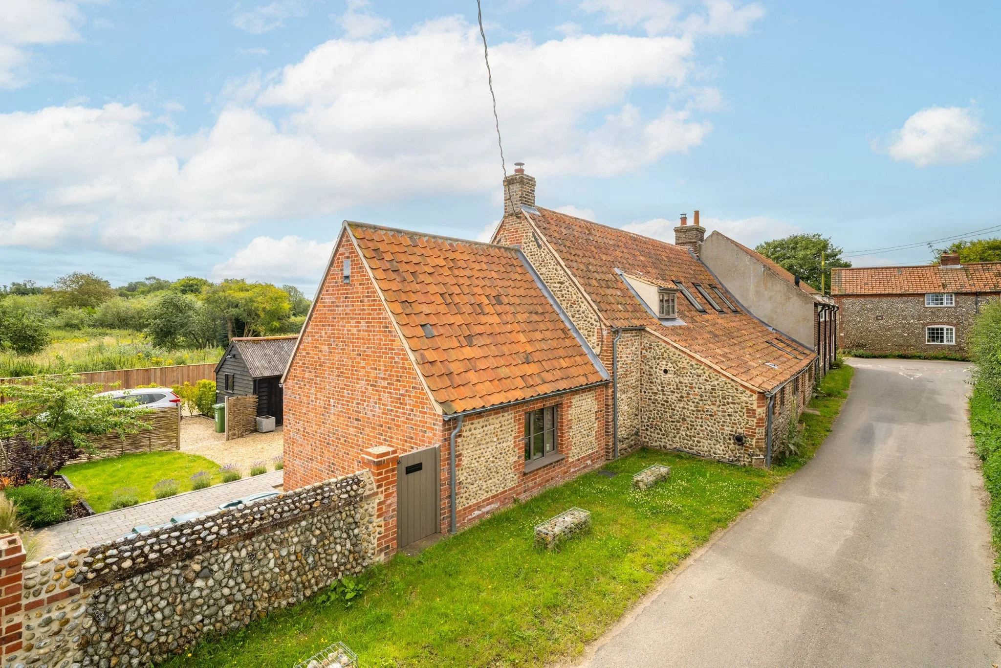 View of a red brick and flint stone house with a tiled roof, chimney, and small windows, situated in a green countryside area with a dirt road.