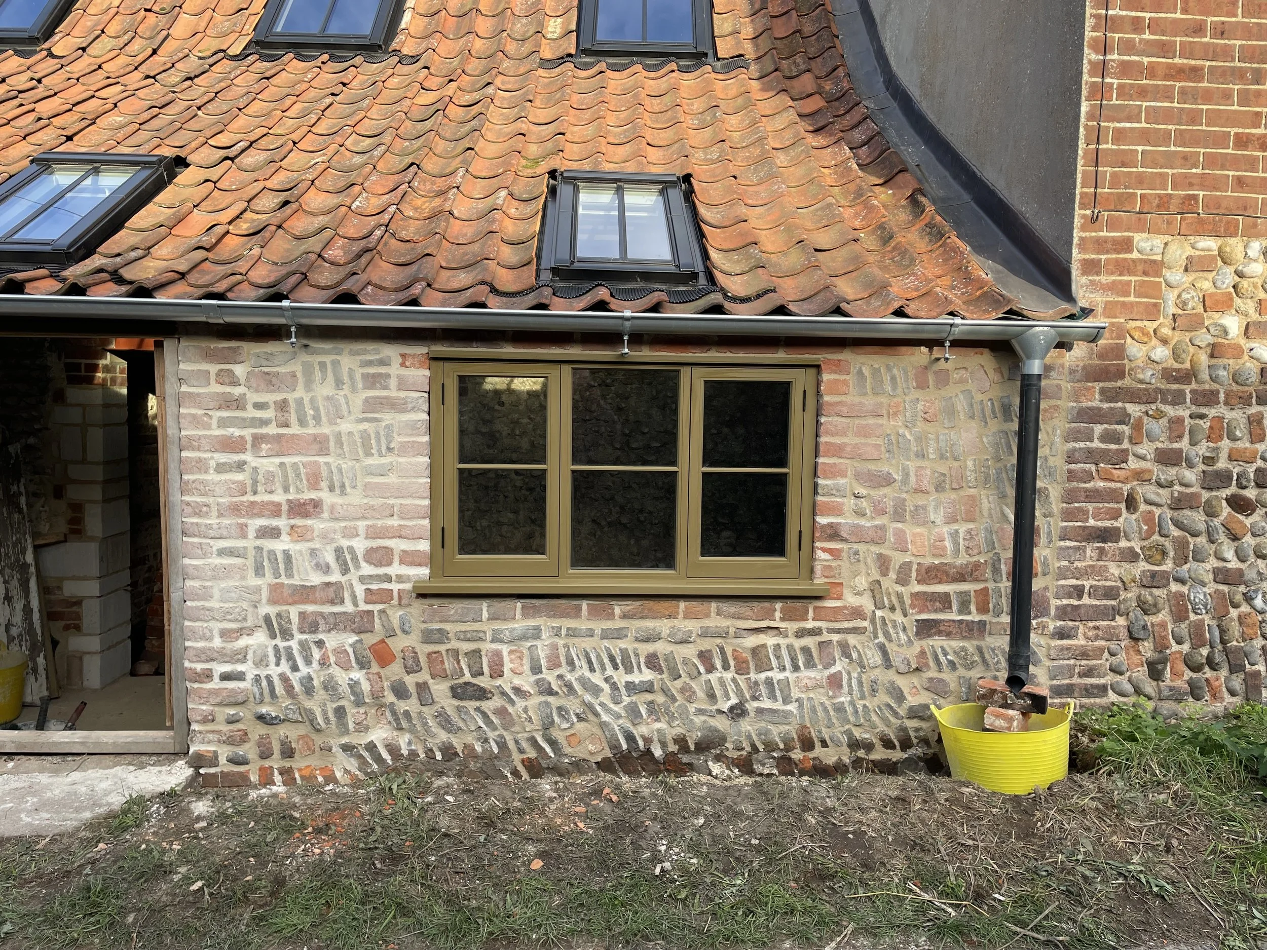A house roof with orange clay tiles, three skylight windows, a brick wall, and a large black downspout pipe. There is also a yellow bucket with bricks and gardening tools outside the house on a grassy and dirt area.