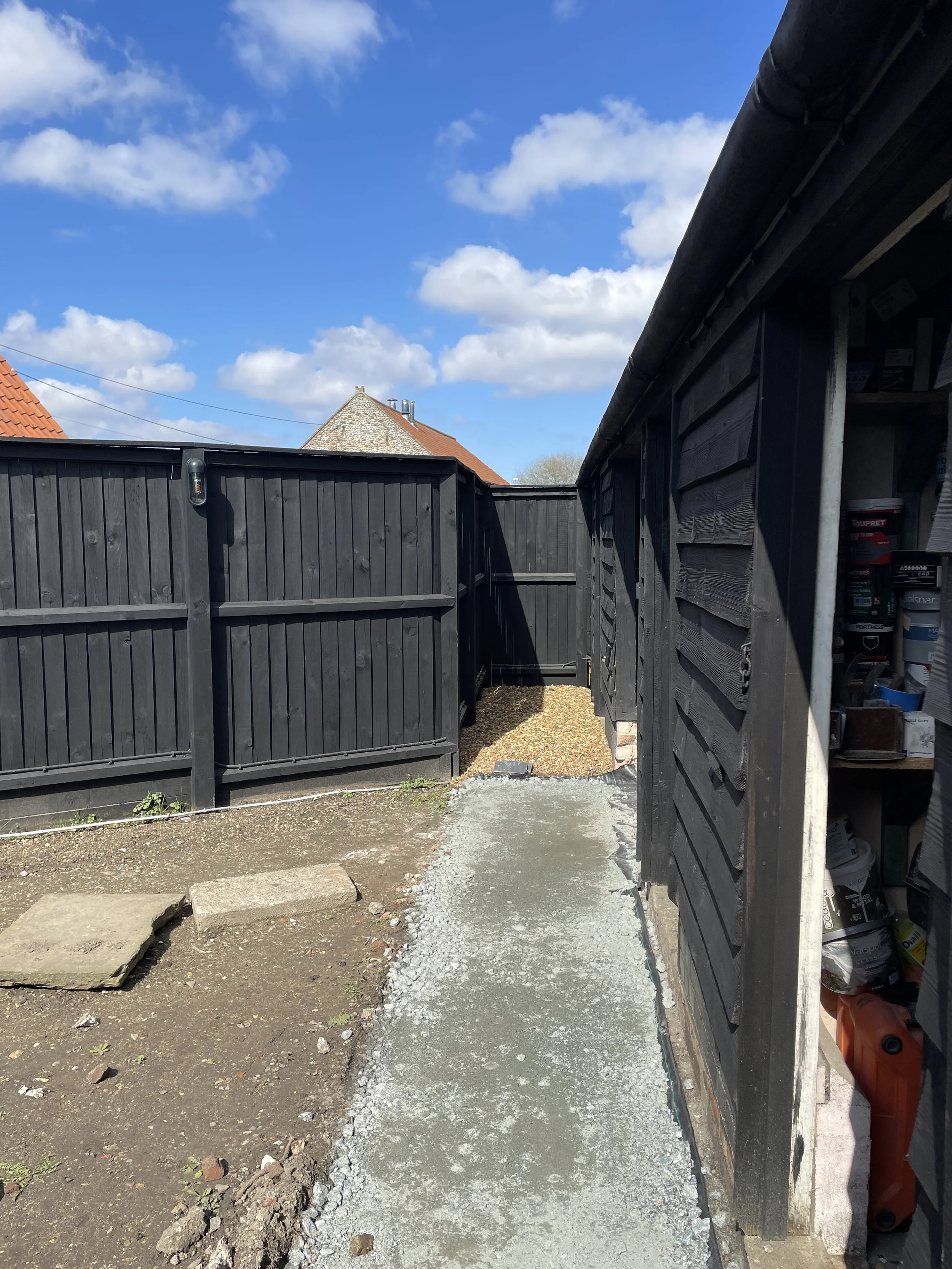 A backyard with a black wooden fence and a black shed on the right, filled with various tools and supplies, and a small gravel pathway under construction, with a gravel and dirt ground, a house with a red roof and a stone chimney in the background, u