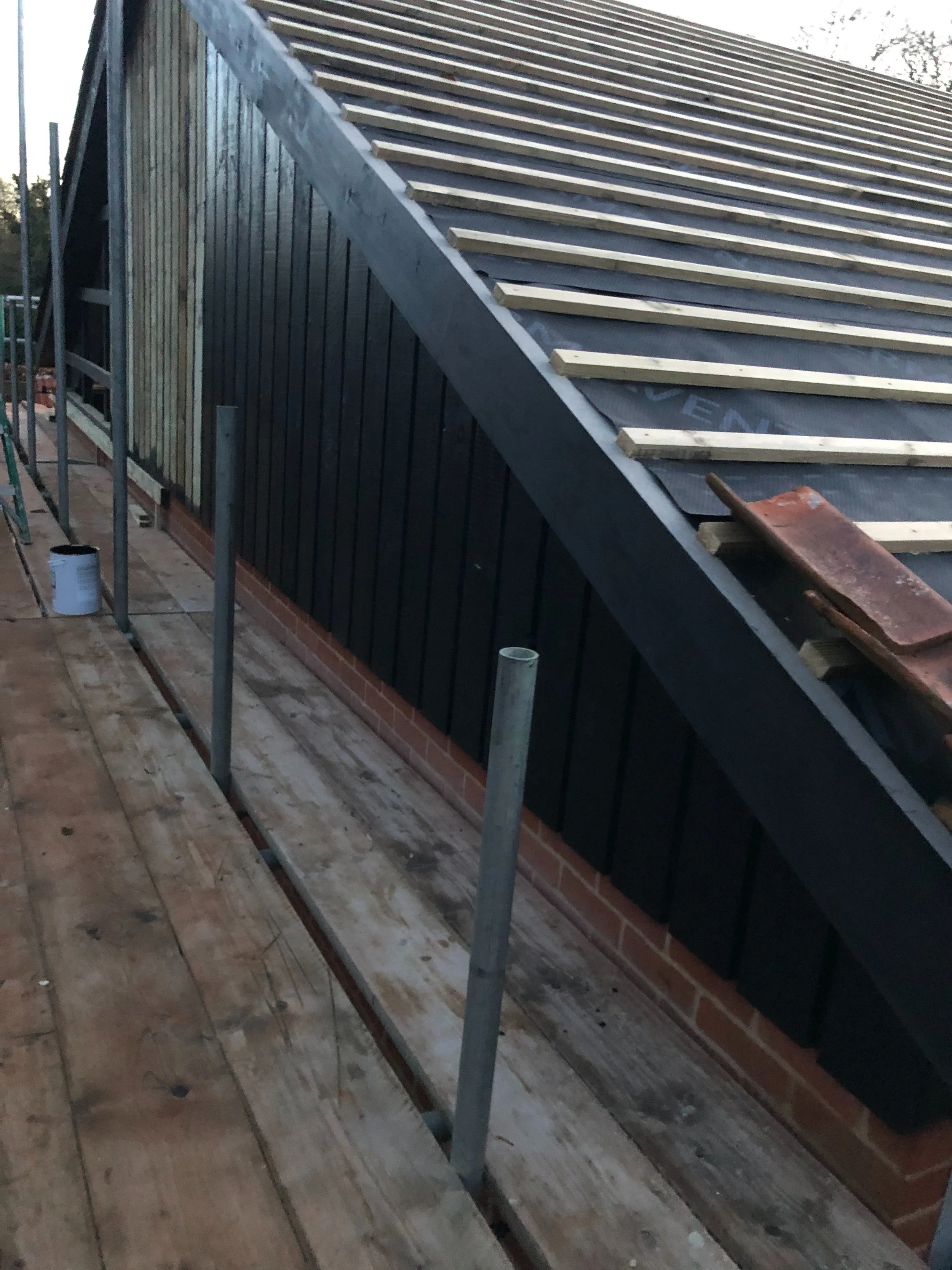 Construction site with a sloped roof under construction, featuring wooden beams and black roofing material. Scaffolding and wooden planks are also visible.