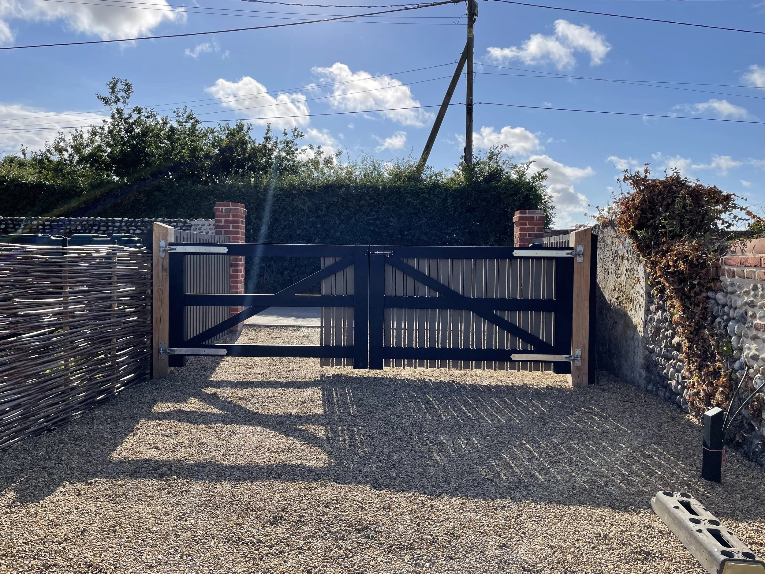 A black metal gate with wooden posts and two brick pillars stands closed in front of a gravel driveway. To the left, there is a woven fence made of natural materials. Behind the gate, a tall hedge and trees extend upward, with a utility pole and powe