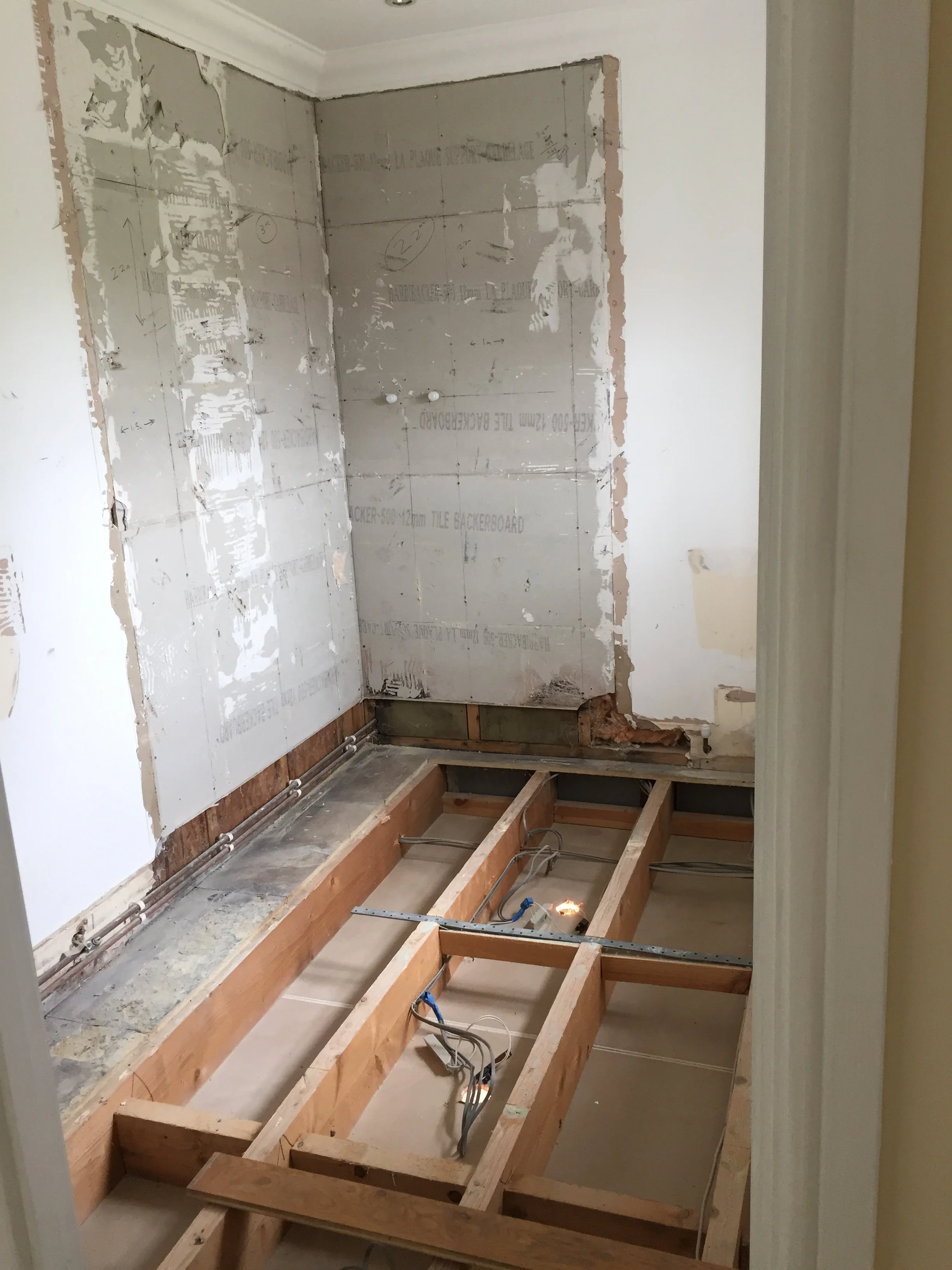 A room under renovation with exposed wooden floor framing, plumbing and electrical wires, and partially removed wall drywall. The floor is unfinished.