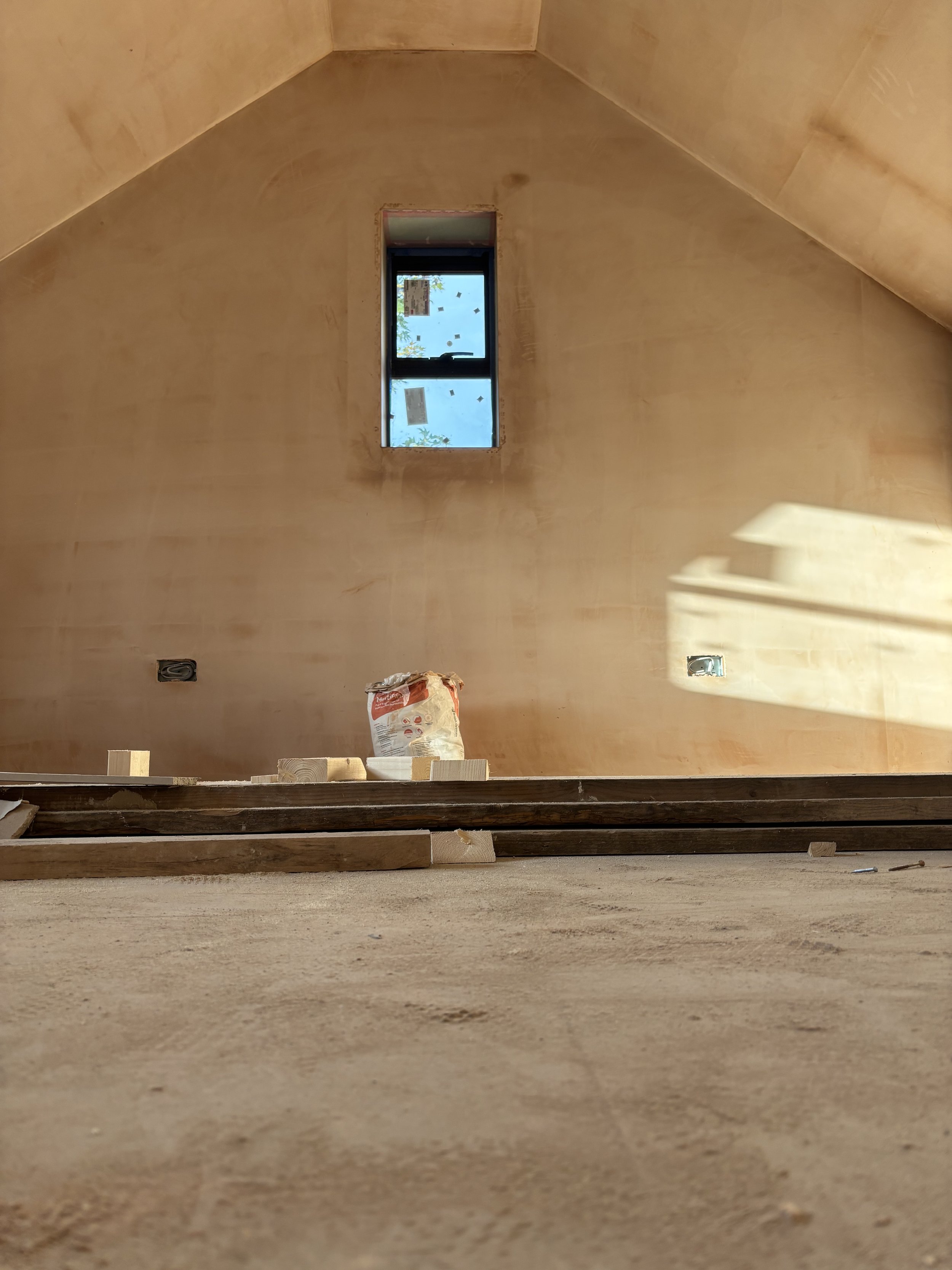 Room under construction with unfinished walls, a small window near the ceiling, and construction materials including a bag of cement and wooden planks.