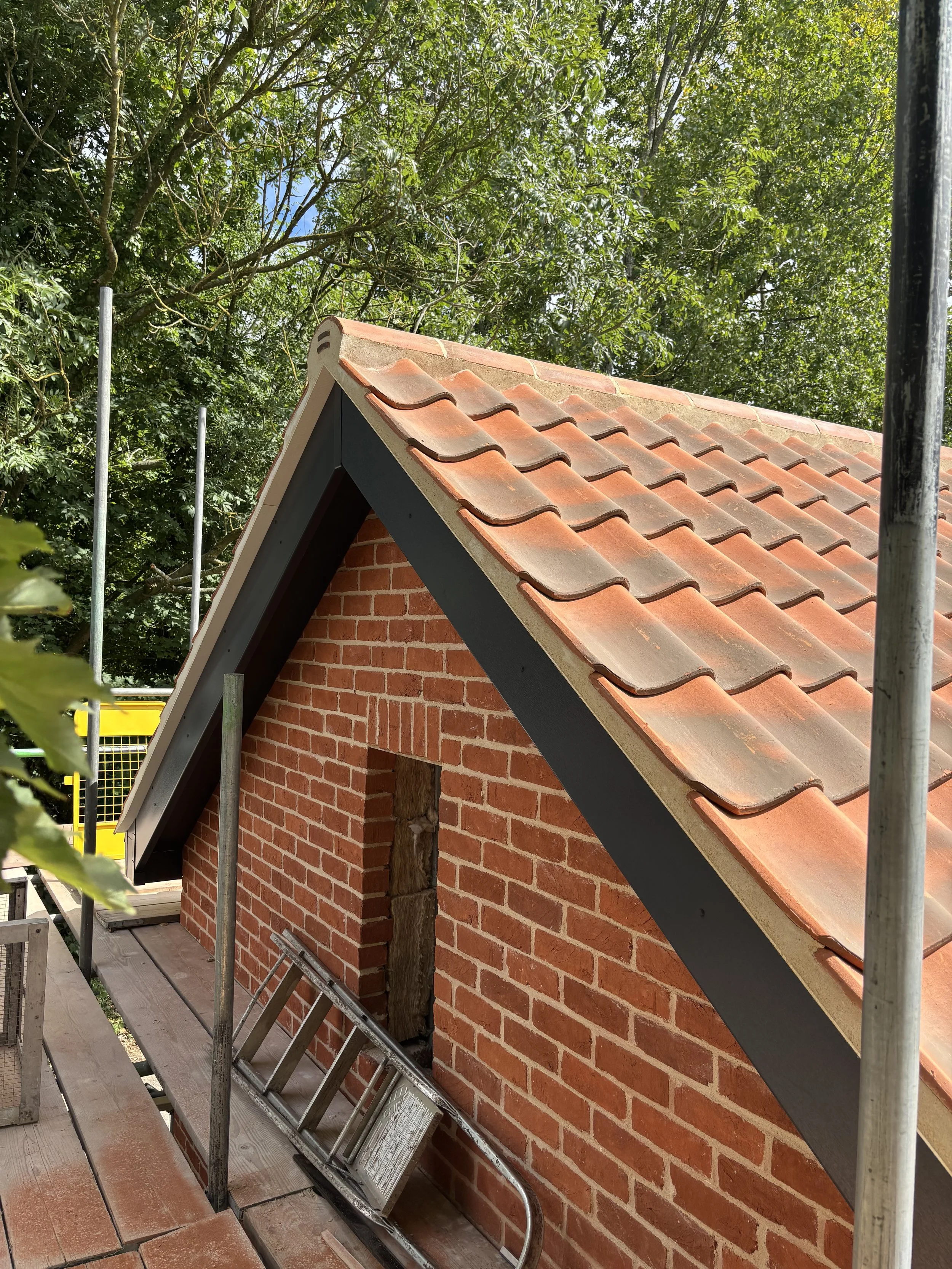 Image showing a house under construction with a brick wall and a pitched roof with red tiles. Scaffolding surrounds the building and a ladder leans against the wall.