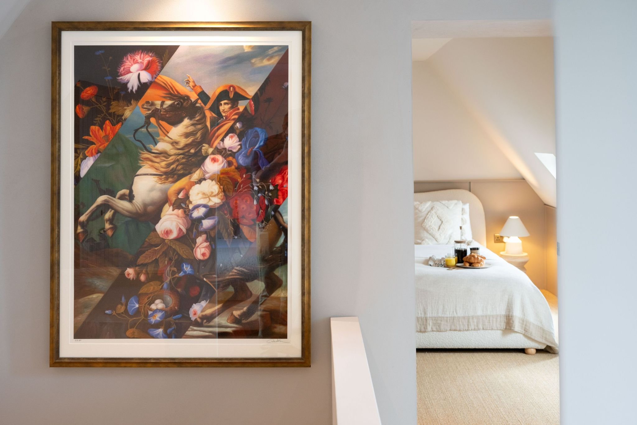 A framed painting of a woman with a bird-shaped hat, surrounded by flowers, hangs on a gray wall in a room with a glimpse of a bed and nightstand with lamp in the background.