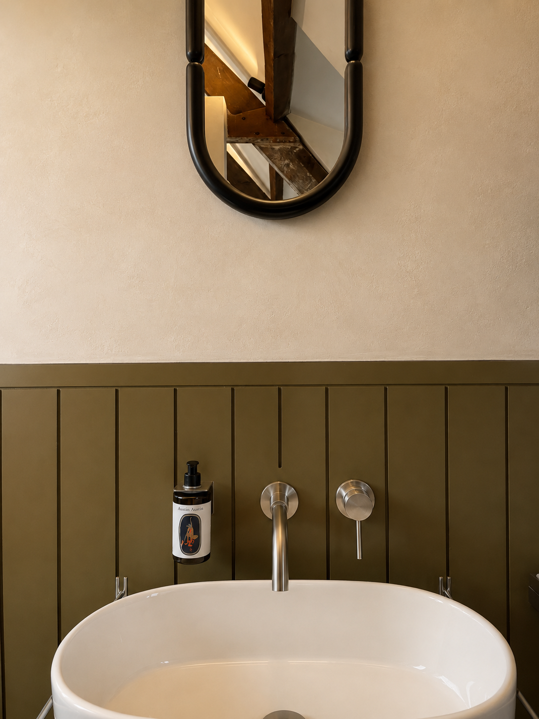 Part of a bathroom with a white oval bathtub, a wall-mounted soap dispenser on the left, and a wall-mounted faucet and handles above the tub. A rectangular mirror with a black frame reflects wooden ceiling beams.