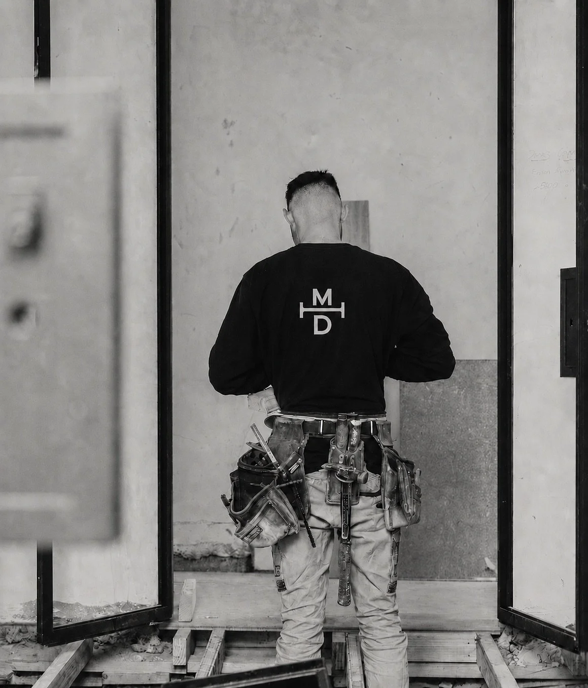 Back view of a construction worker standing on a wooden platform, wearing a tool belt and a jacket with initials, inside a building under construction.