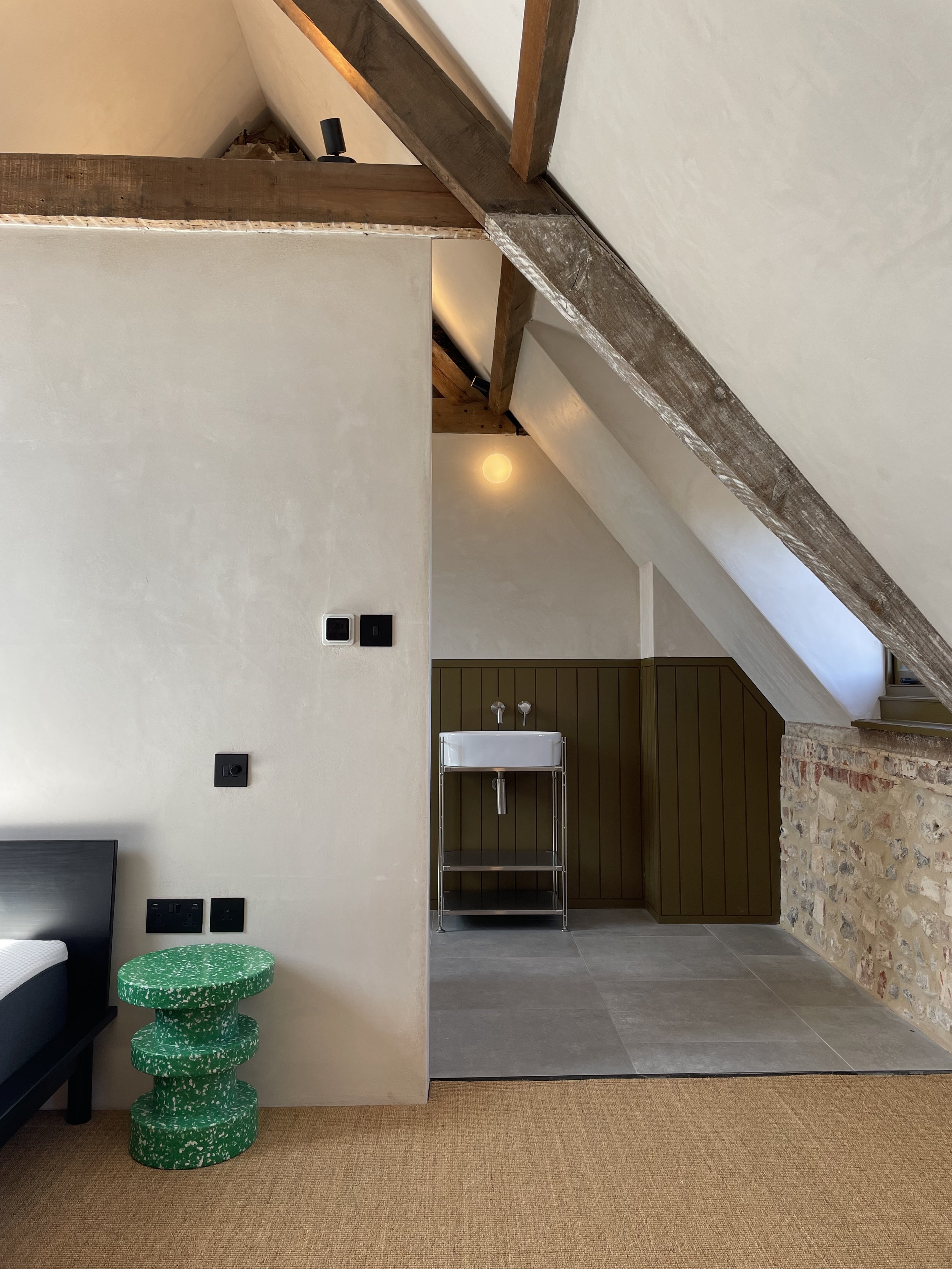 Interior view of a room with a sloped ceiling, wooden beams, a small wash basin, and a green, stepped side table. The wall has a textured, exposed brick section near the floor, with a beige carpet covering part of the floor.