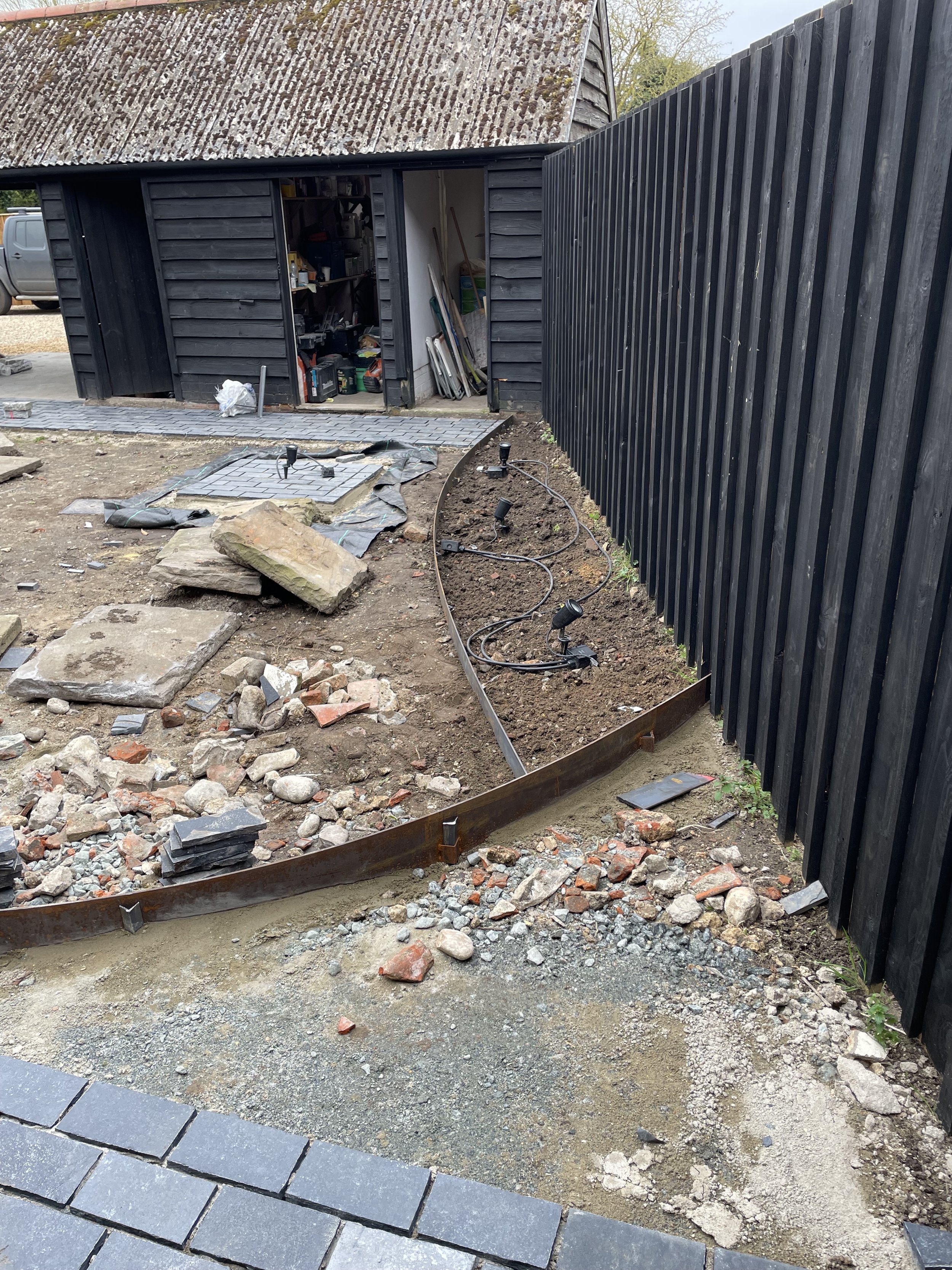 A garden or yard area under construction with an unfinished pathway. There are paving stones on the ground, a curved metal edge, dirt, rocks, and construction tools. A black wooden shed with shelves is open, and there is a black fence on one side.