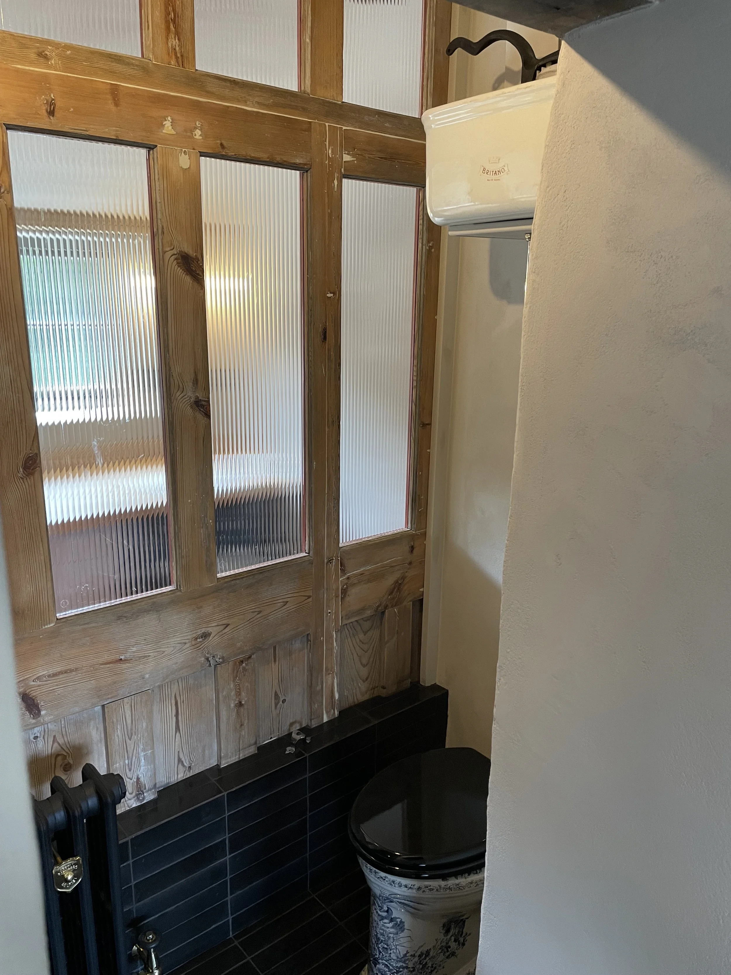 Bathroom with a wooden door featuring frosted glass panels, black tiled floor, a white toilet with decorative blue designs, and a small white cabinet with a curved top.